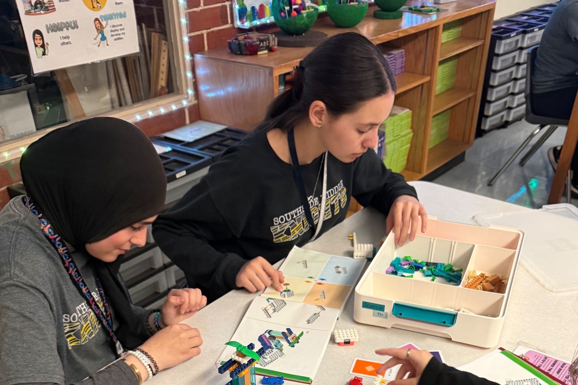 Three women at a table work on a craft project, with colorful beads, scissors, and a pink backpack.