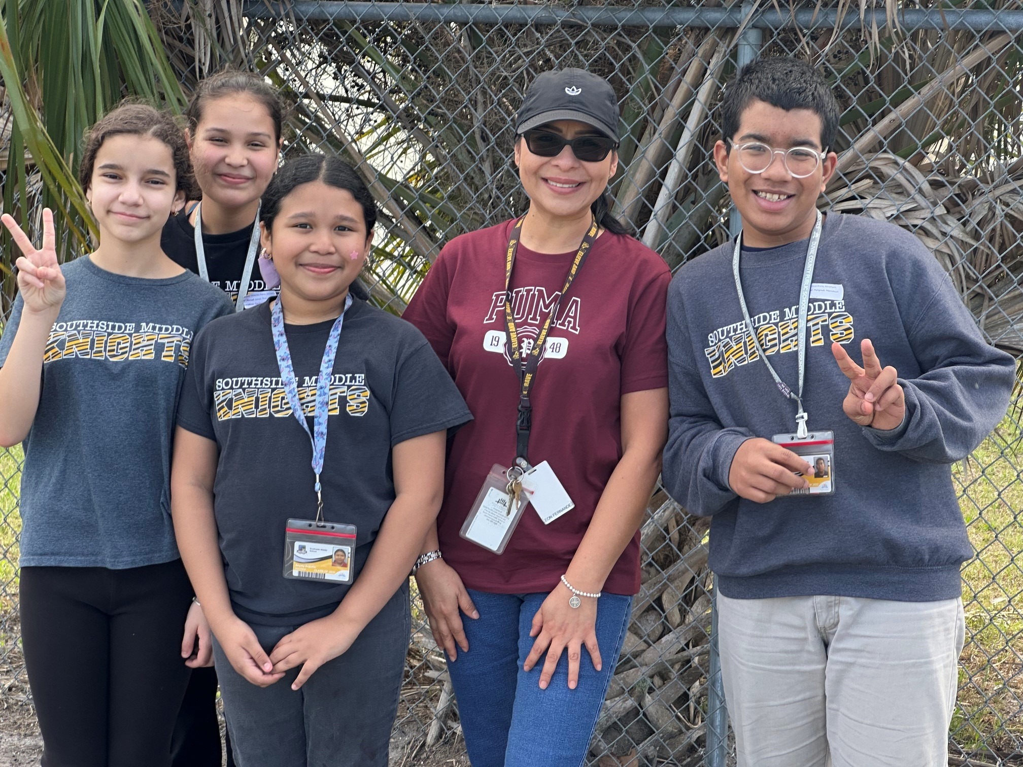Five people stand in front of a fence, all wearing ID lanyards. They pose for a photo, making peace signs.