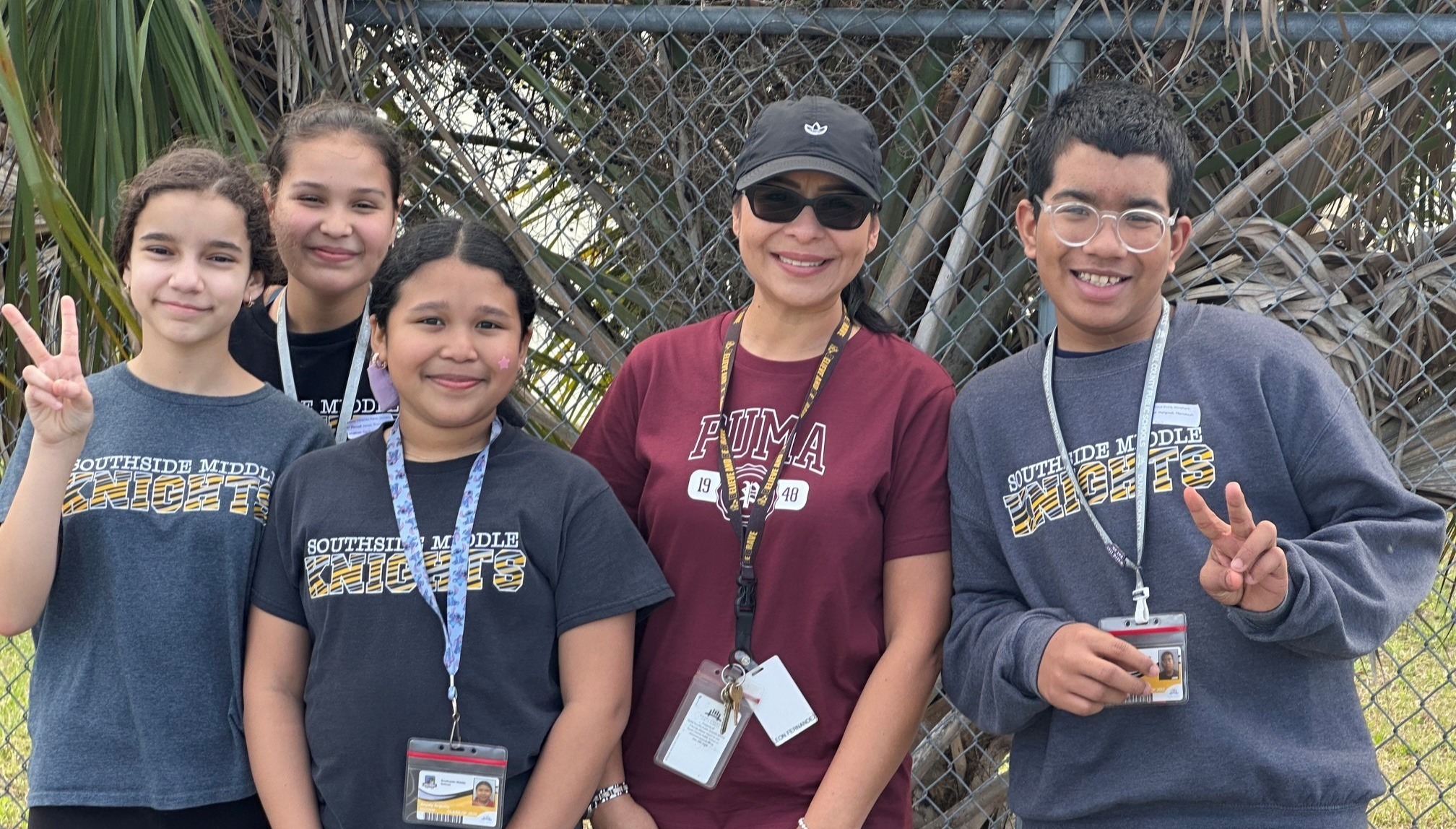 Five people stand in front of a fence, all wearing ID lanyards. They pose for a photo, making peace signs.