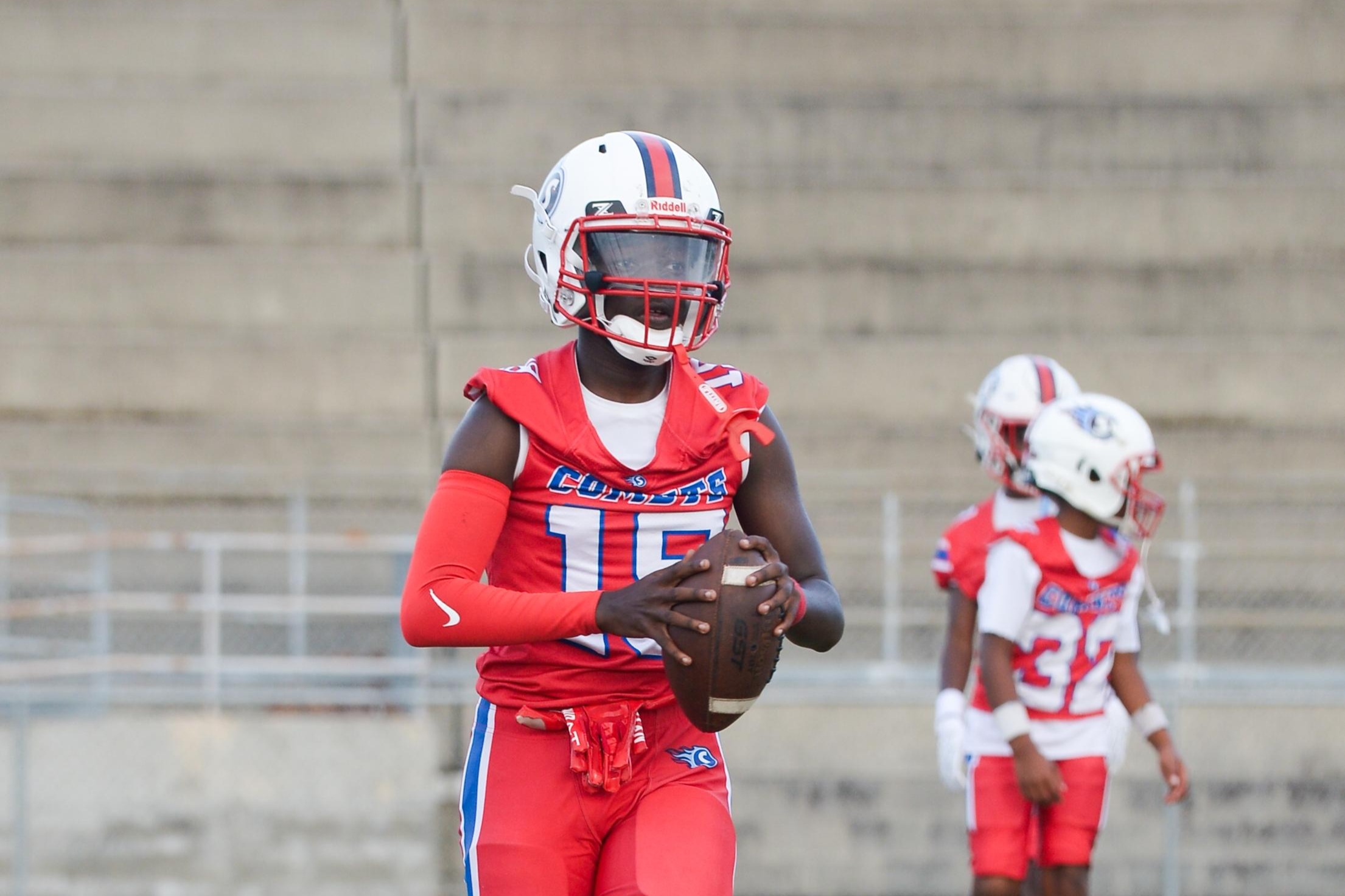 A football player in red jersey and white helmet holds a ball on a grassy field with blurred players in the background.