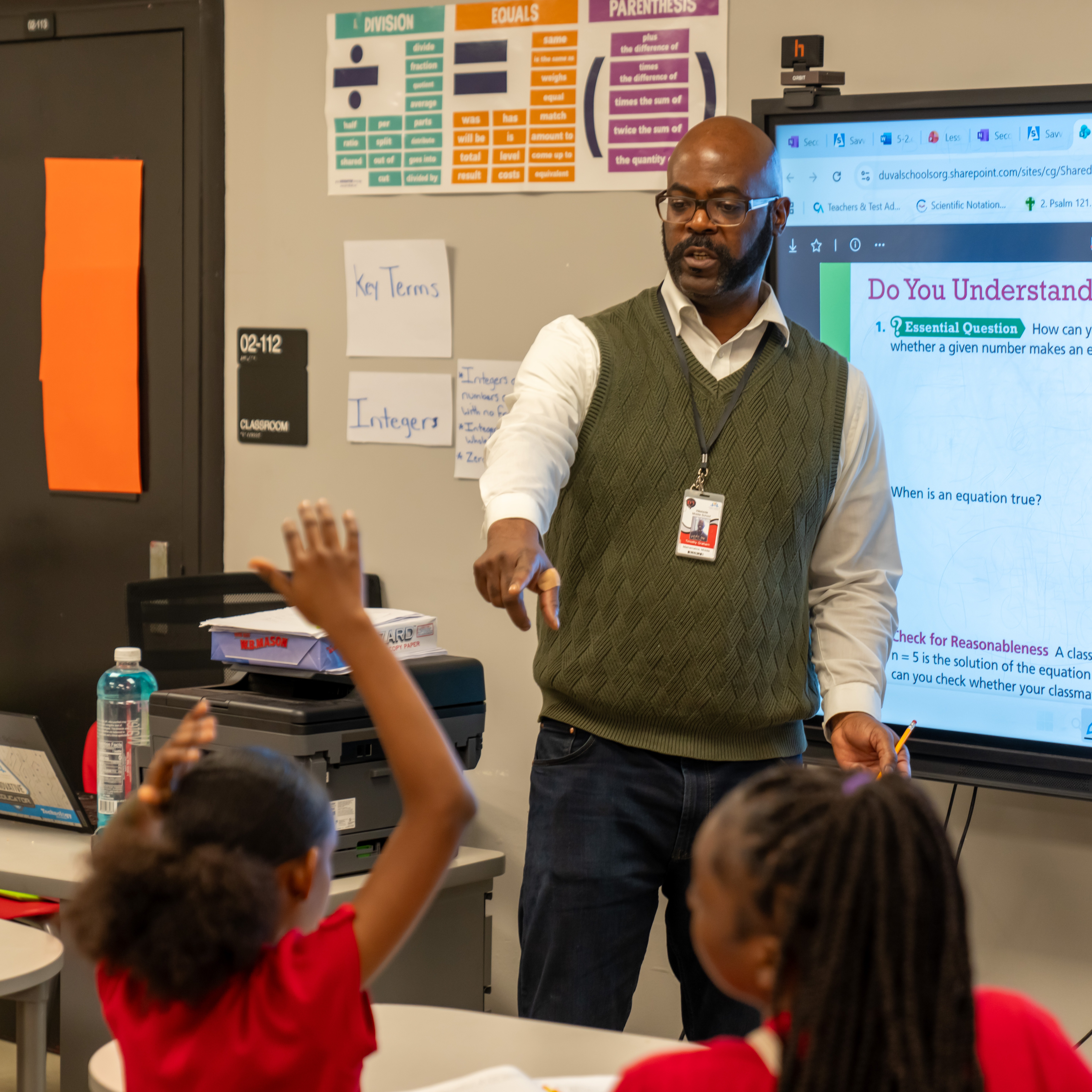 A teacher in front of a screen displays a math problem. Two students raise their hands.