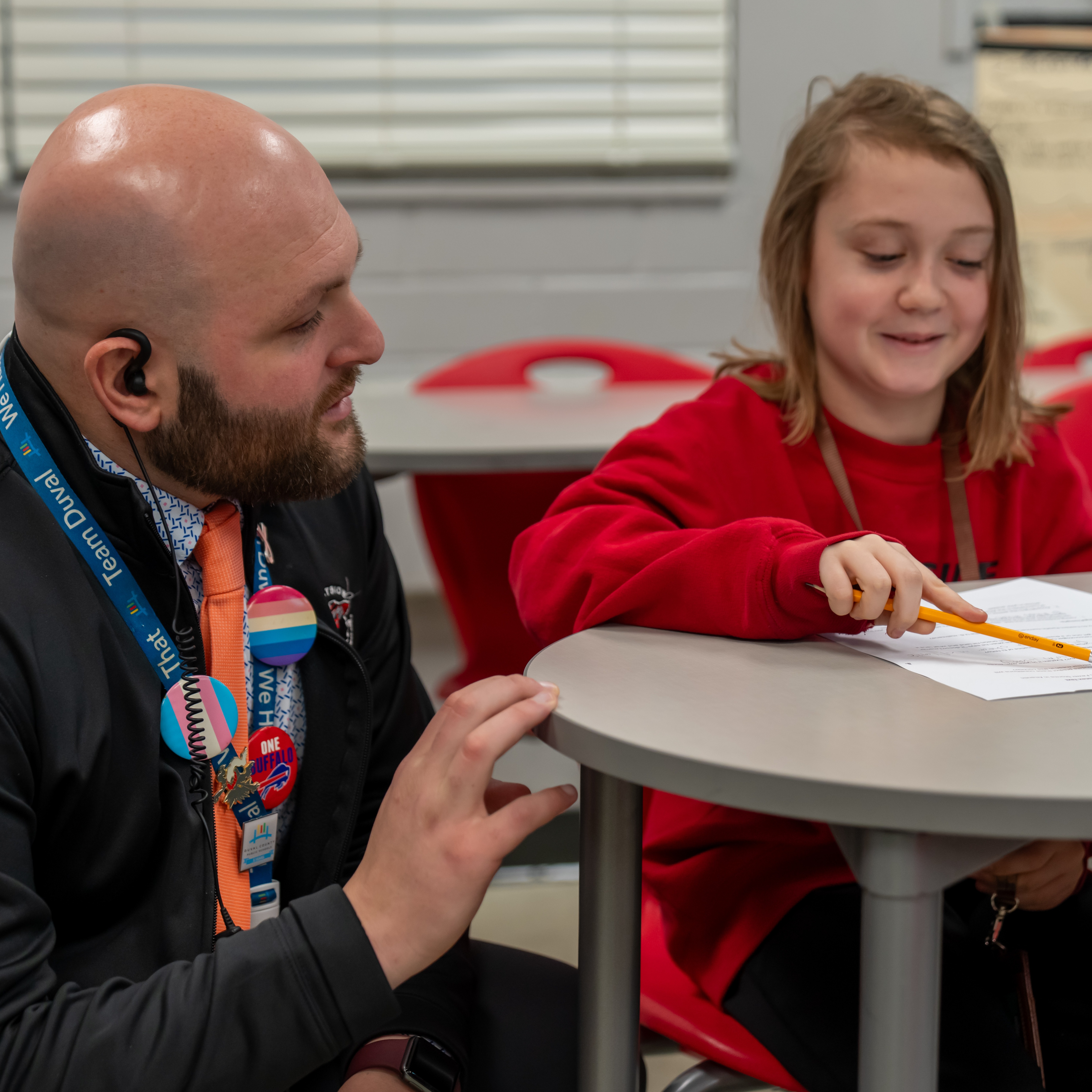 A man with a badge sits next to a girl, who is smiling while writing.
