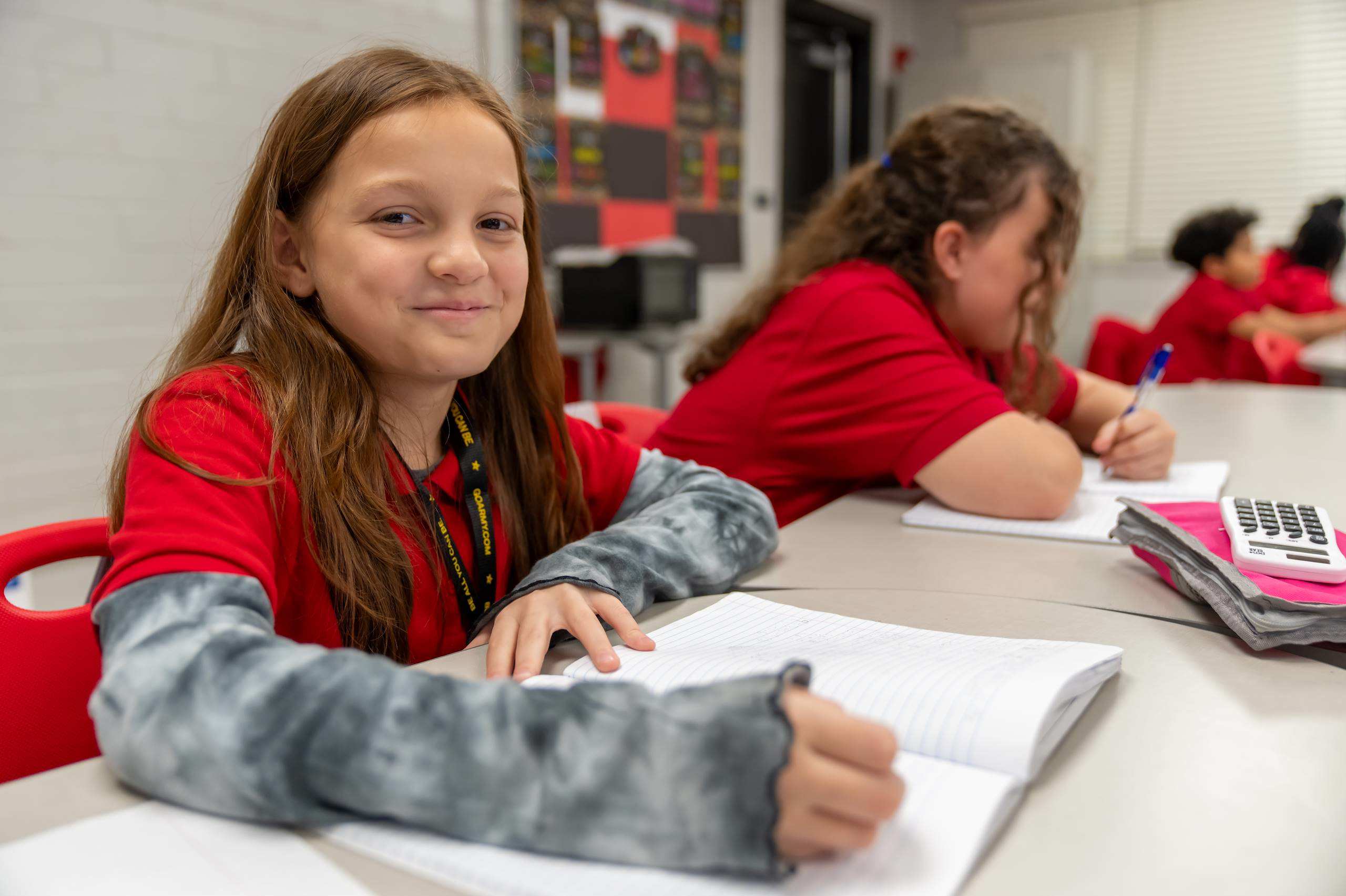 A young girl in a red shirt sits at a table, smiling, holding a pen and notebook. Other students, also in red shirts, are seated nearby, working on their papers.