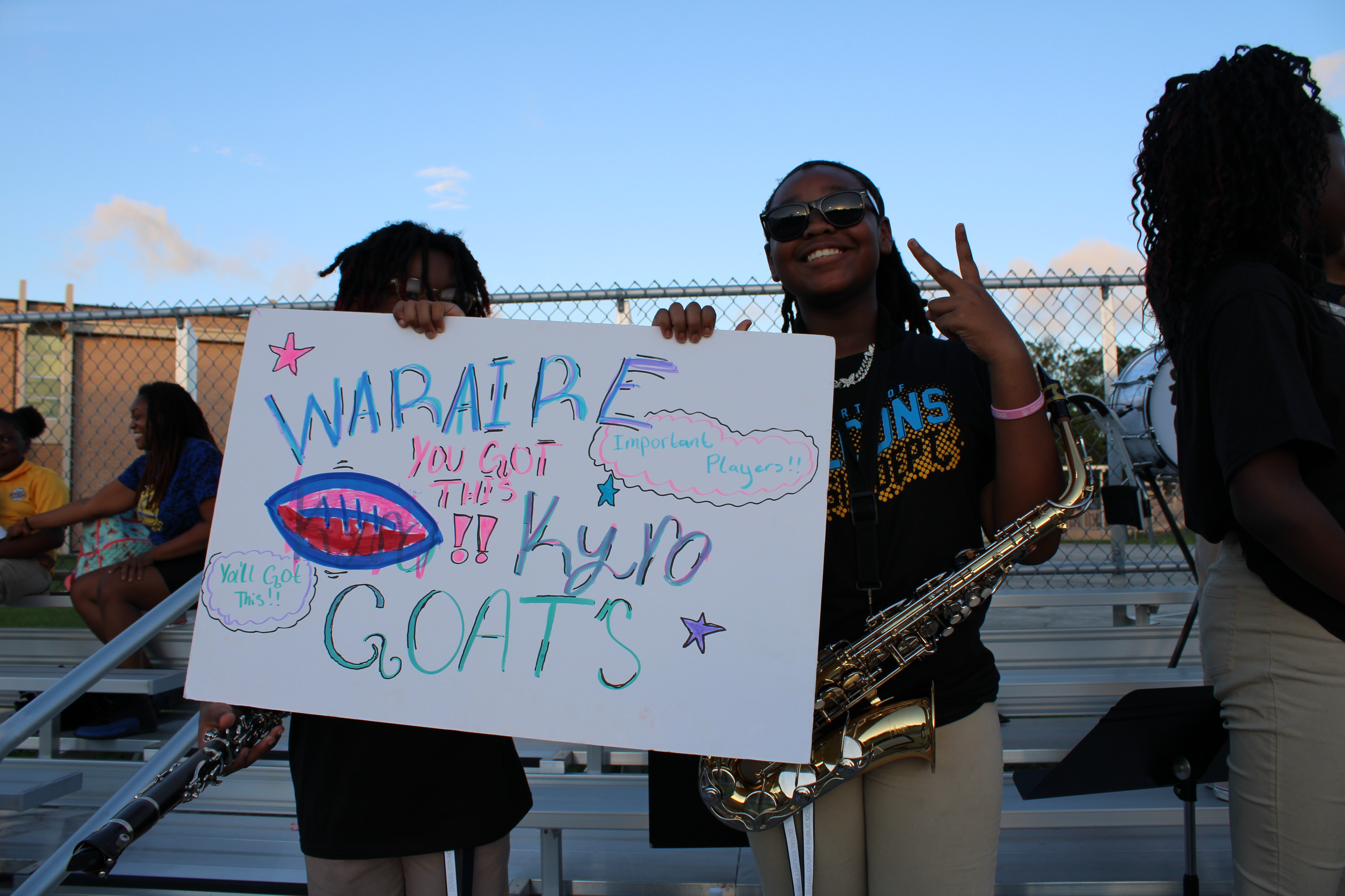 Two women hold a sign that says "WARAIRE YOU CAN'T STOP THE KYRO GOATS". One is holding a saxophone.