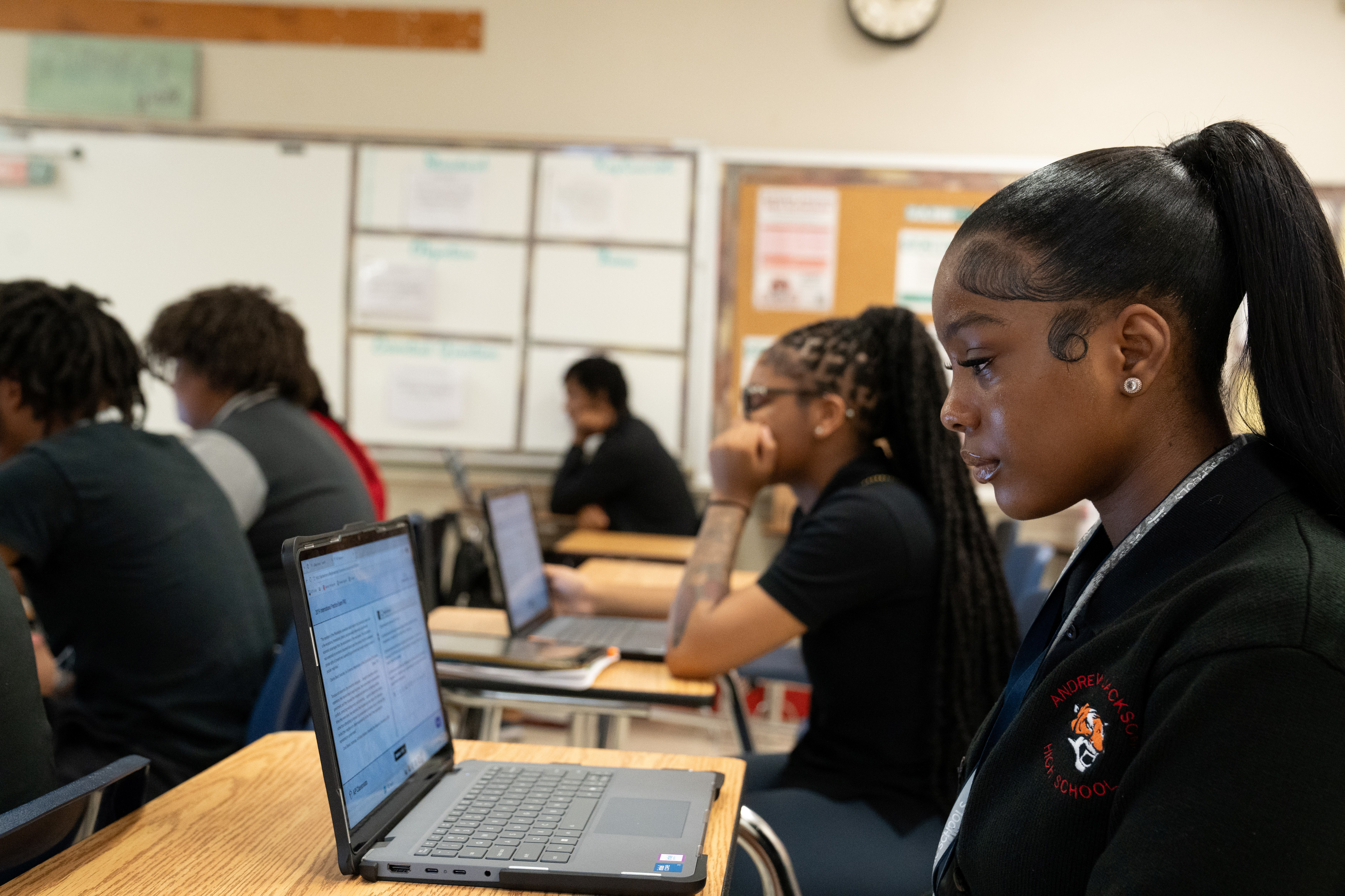 Students in a classroom, some seated and using laptops on their desks, while a woman looks at a laptop screen.