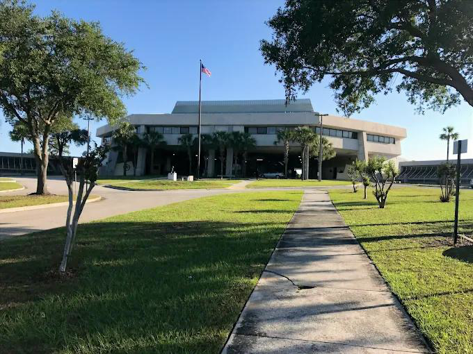 Pathway leads to a large building with a flagpole, set on a grassy lawn with trees.