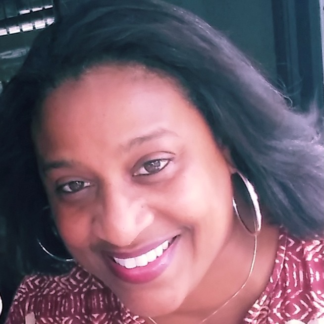 Woman with dark hair wearing a red and white patterned shirt, smiling, and sporting hoop earrings.