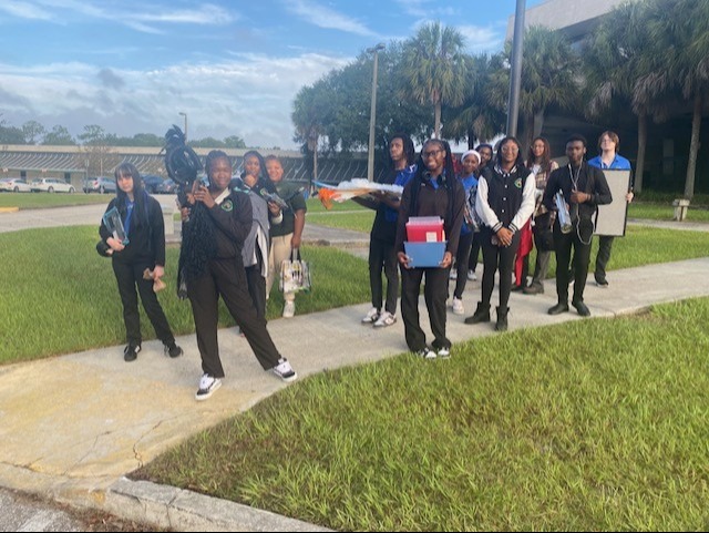A group of people dressed in matching uniforms walk on a sidewalk. Some hold items and a building is visible in the background.