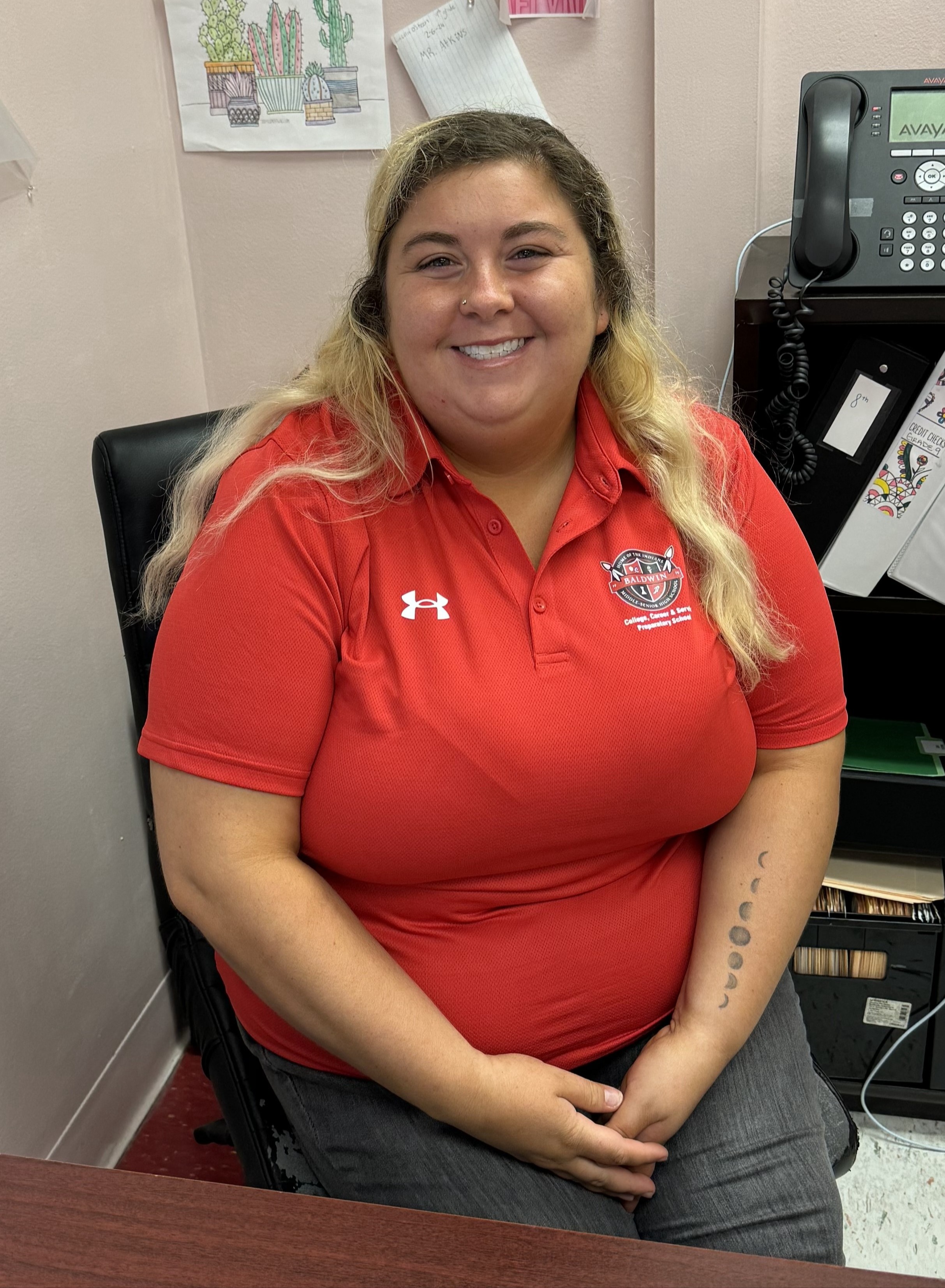 A woman in a red polo sits in a chair, smiling, with a pink wall behind her. A telephone is to the right.