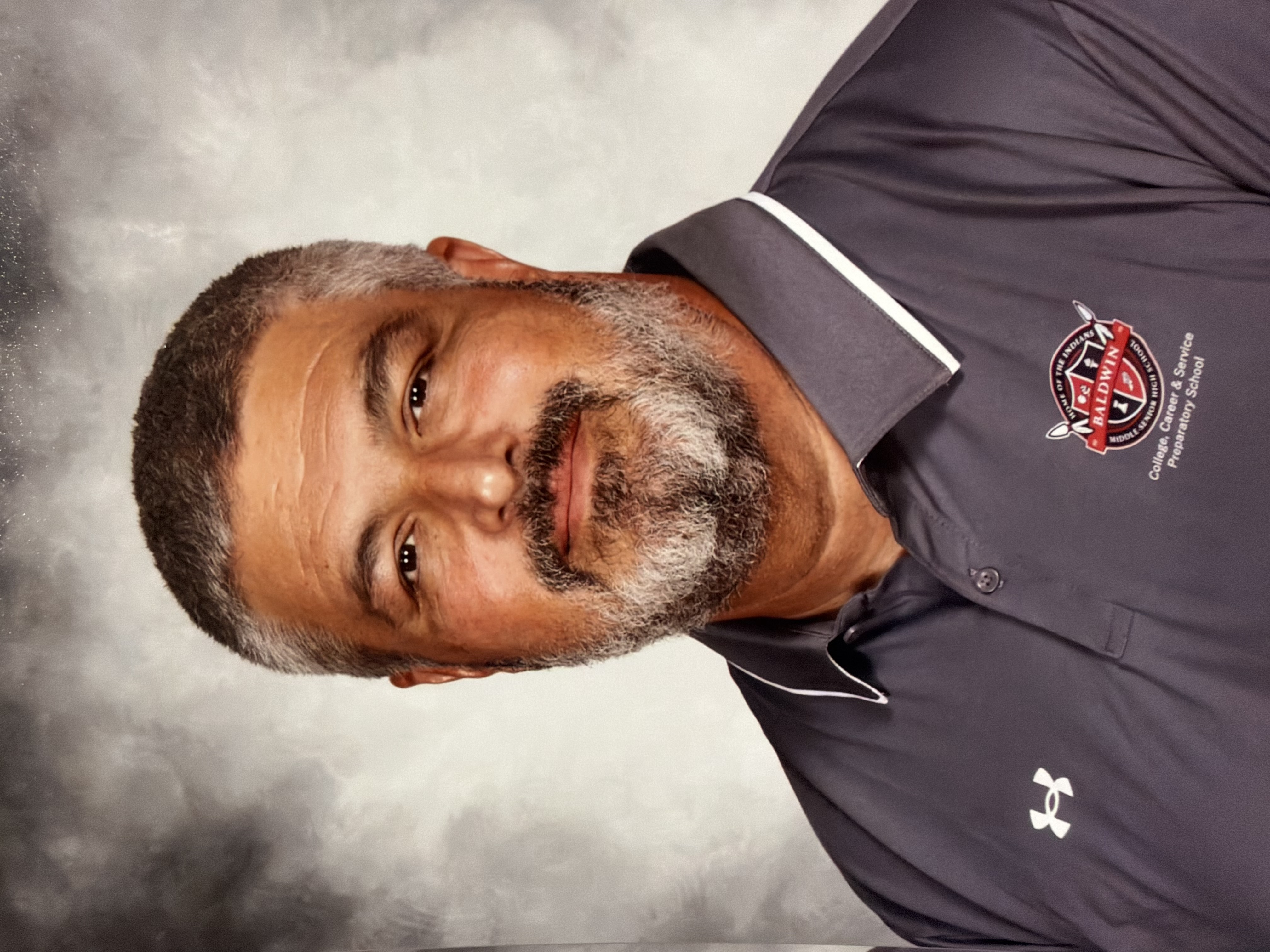 A man with a beard, wearing a gray Under Armour polo, is smiling and looking at the camera.