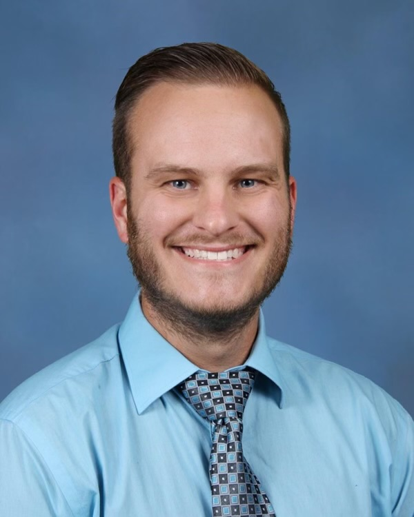 A man with a beard and short hair in a blue shirt and tie. He is smiling.