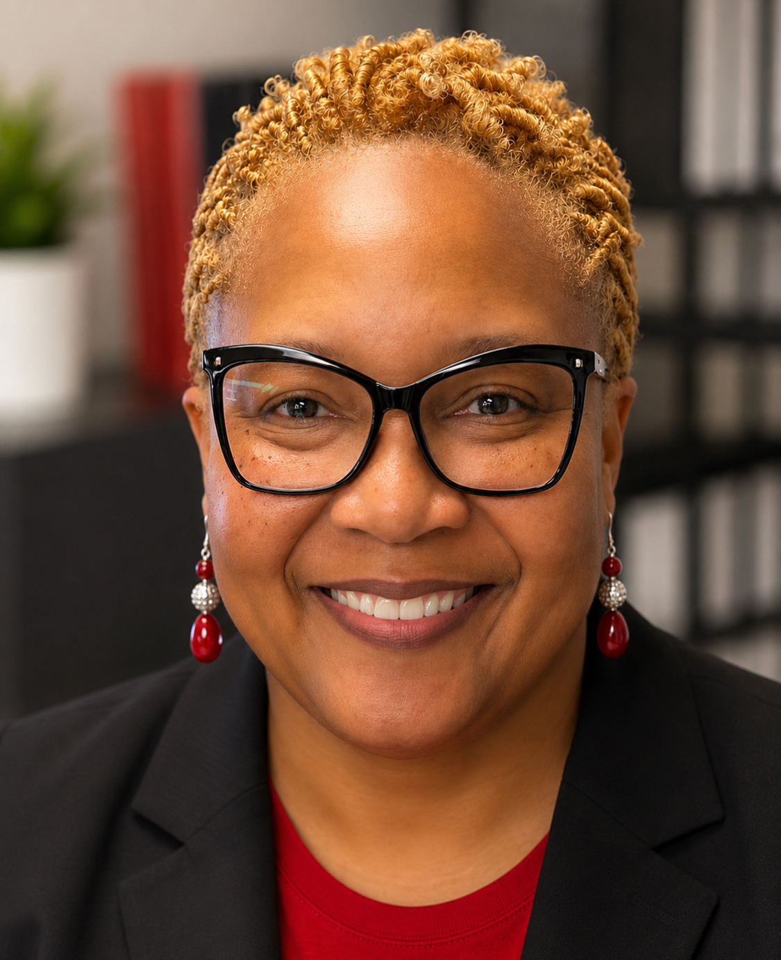 Woman with short hair and glasses, wearing black blazer, red shirt, and earrings, smiles for the camera.