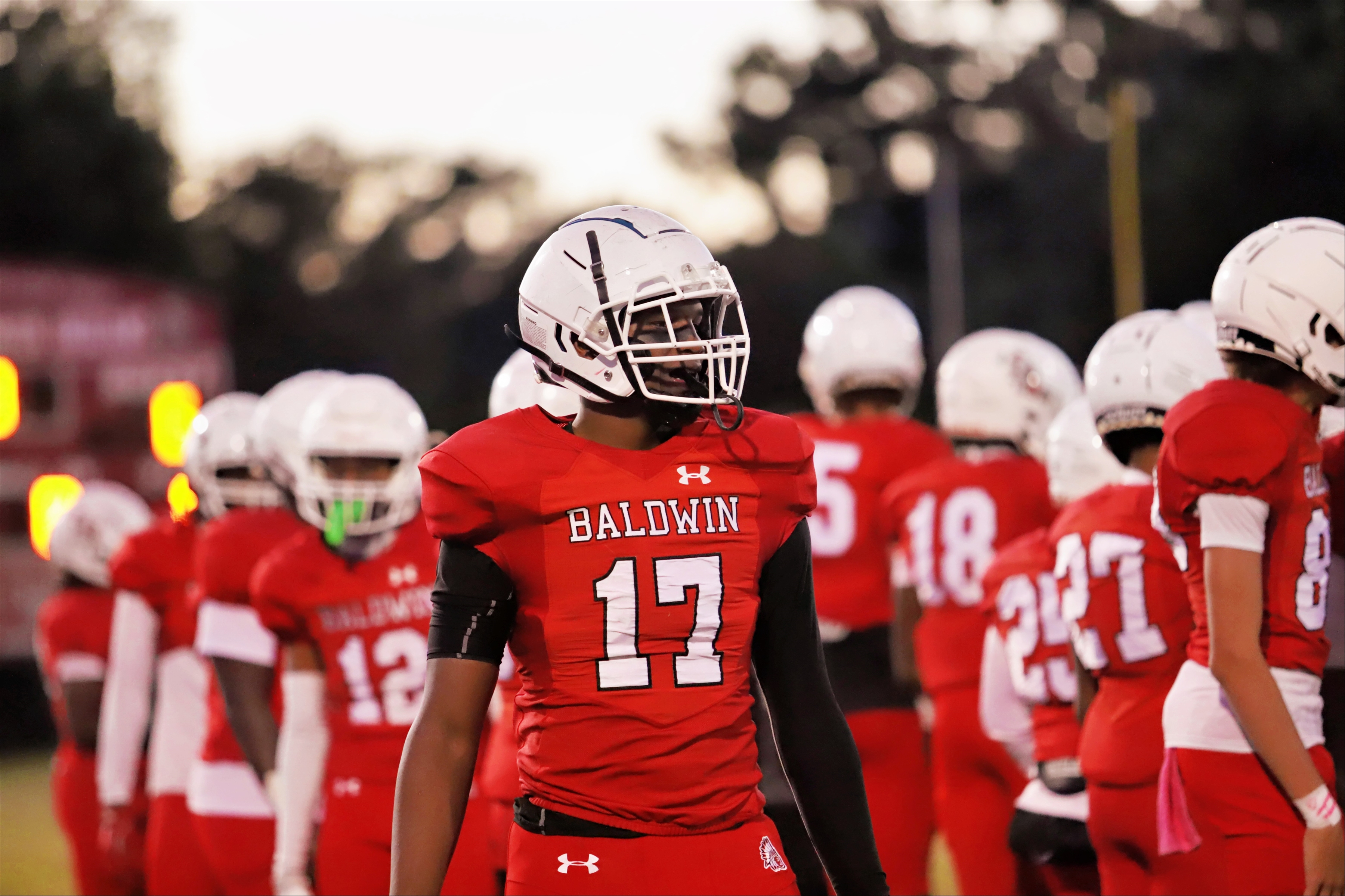 Football players wearing red uniforms and white helmets are lined up on the field. One player wears a red jersey with number 17.