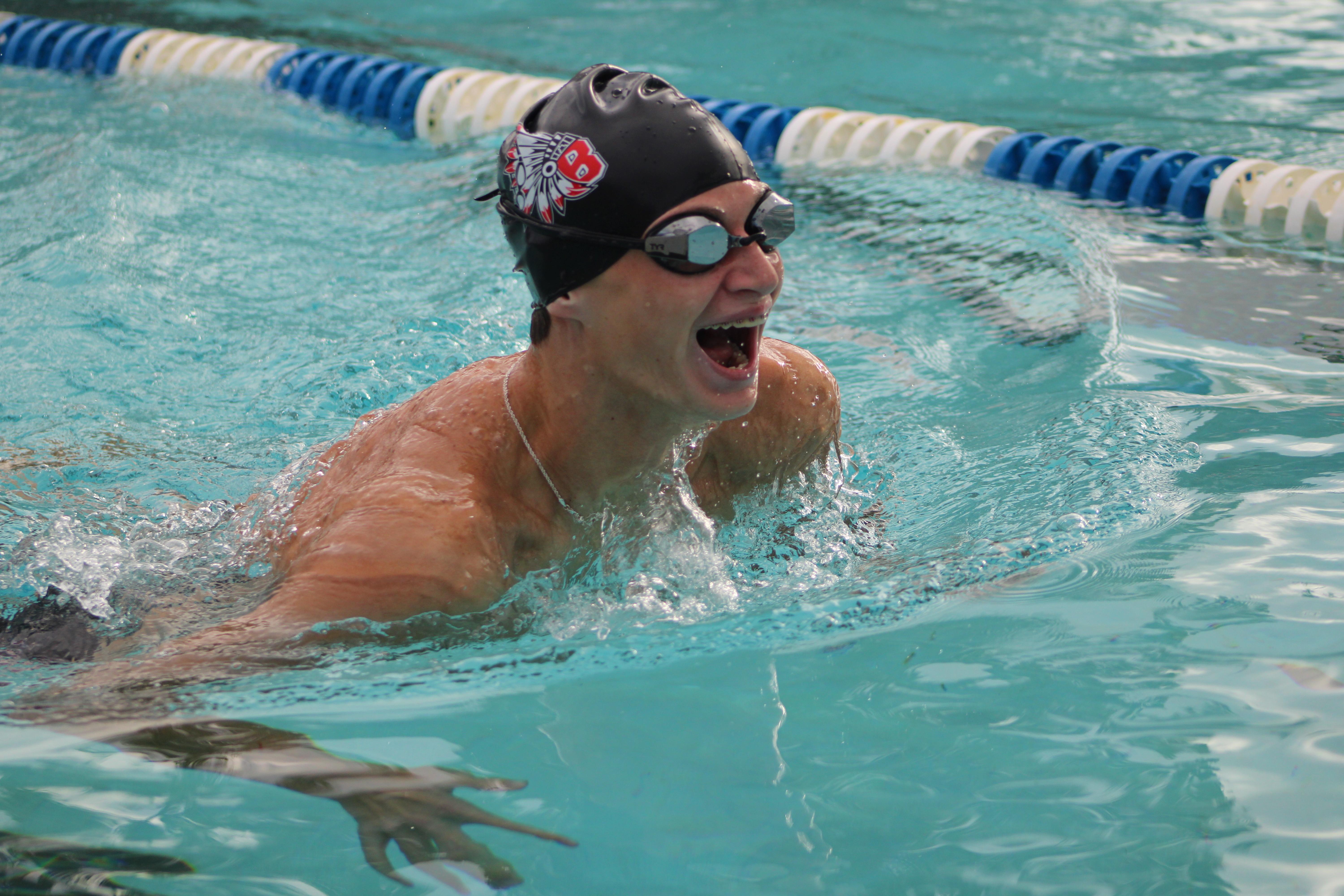A man swimming in a pool wearing a swim cap and goggles, his mouth open as if shouting.