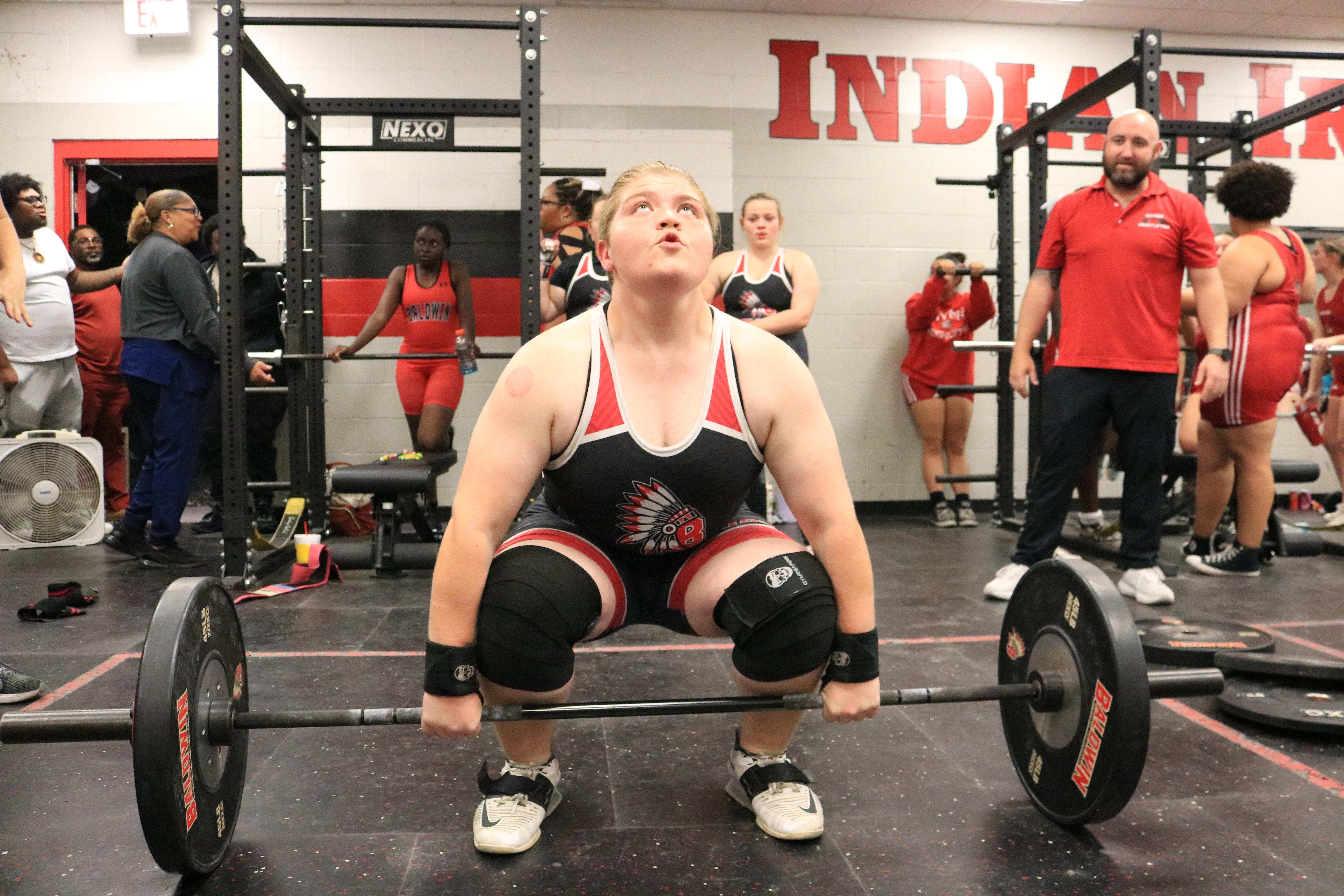 An athlete lifts a barbell in a gym, wearing a red and white uniform. Several people watch in the background.