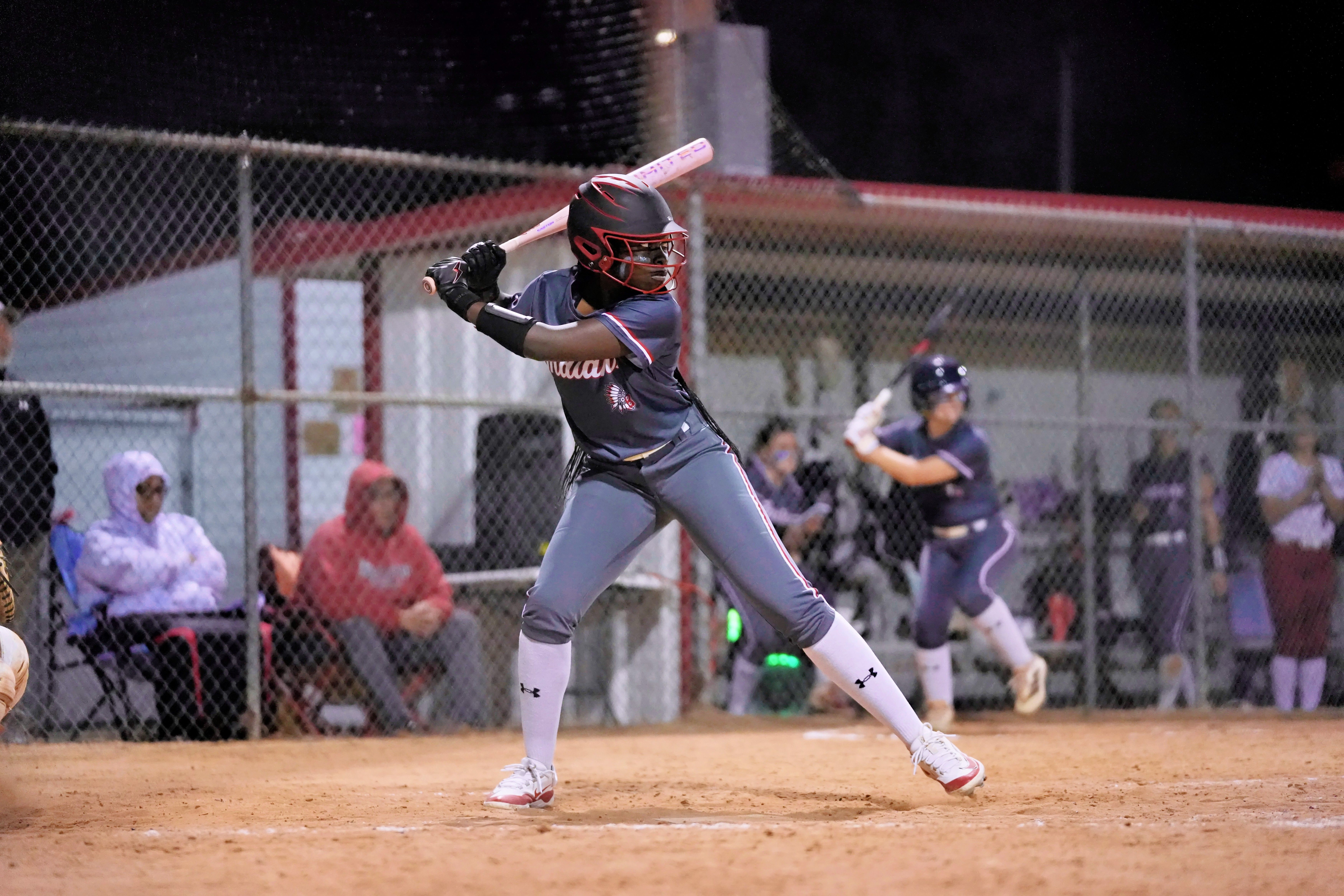 A baseball player swings a bat on a field, with a blurred view of spectators and another player in the background.
