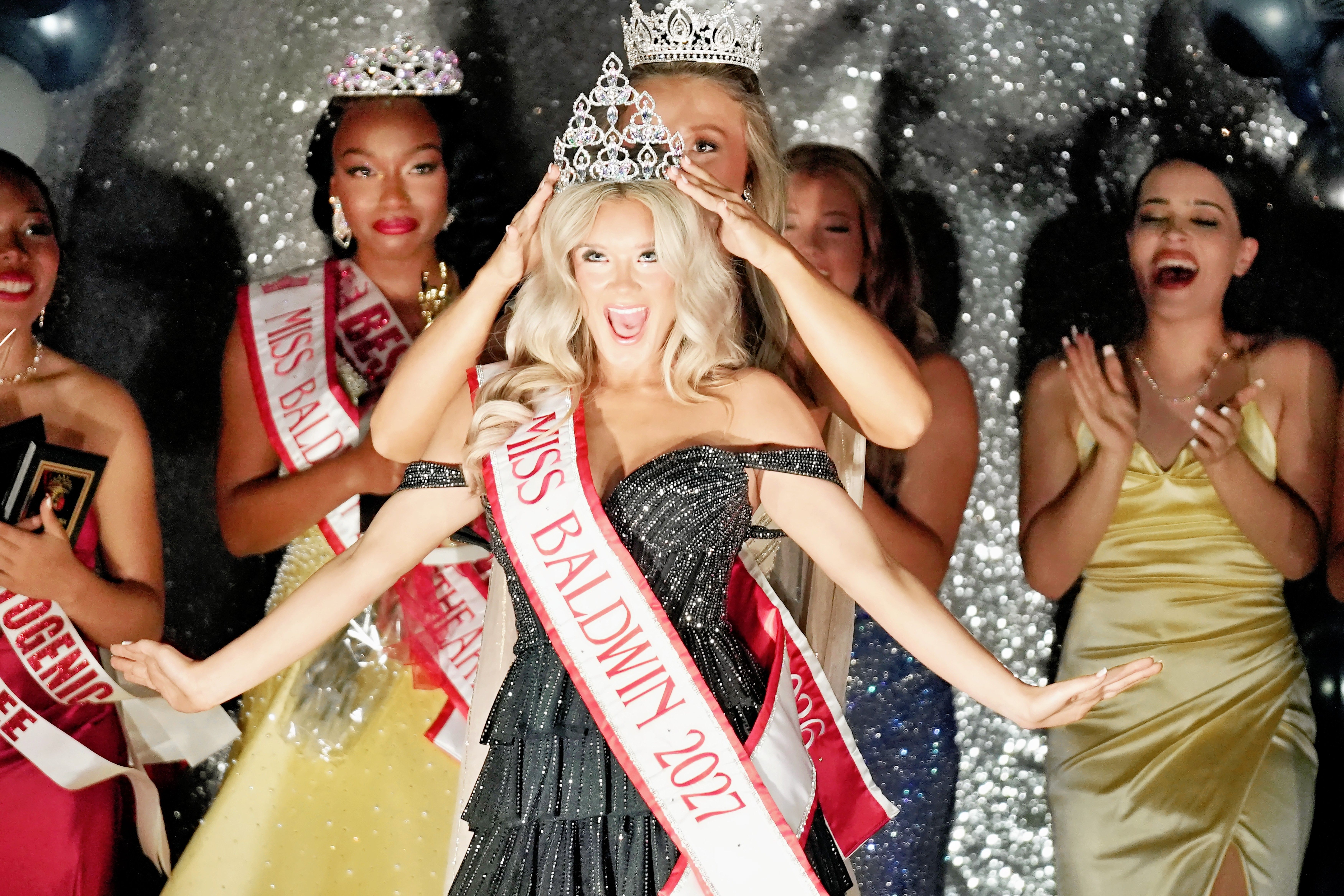 Contestants in gowns and sashes celebrate in a room with glitter and balloons in the background.