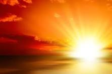 Bright sun setting on horizon, illuminating sky with warm orange and yellow hues, clouds partly covering the sun.