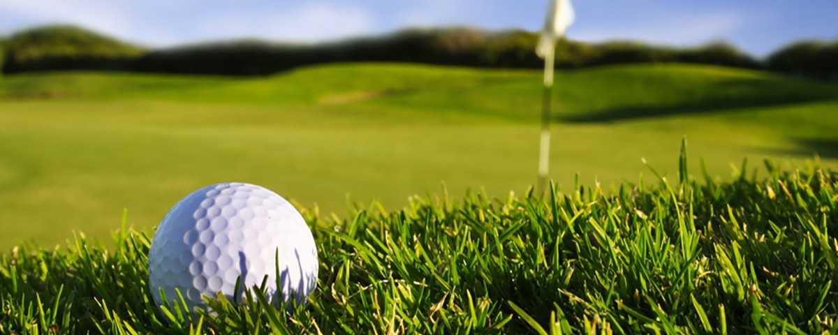 A white golf ball is on the grass near a white flagstick. The background is a golf course with rolling hills.