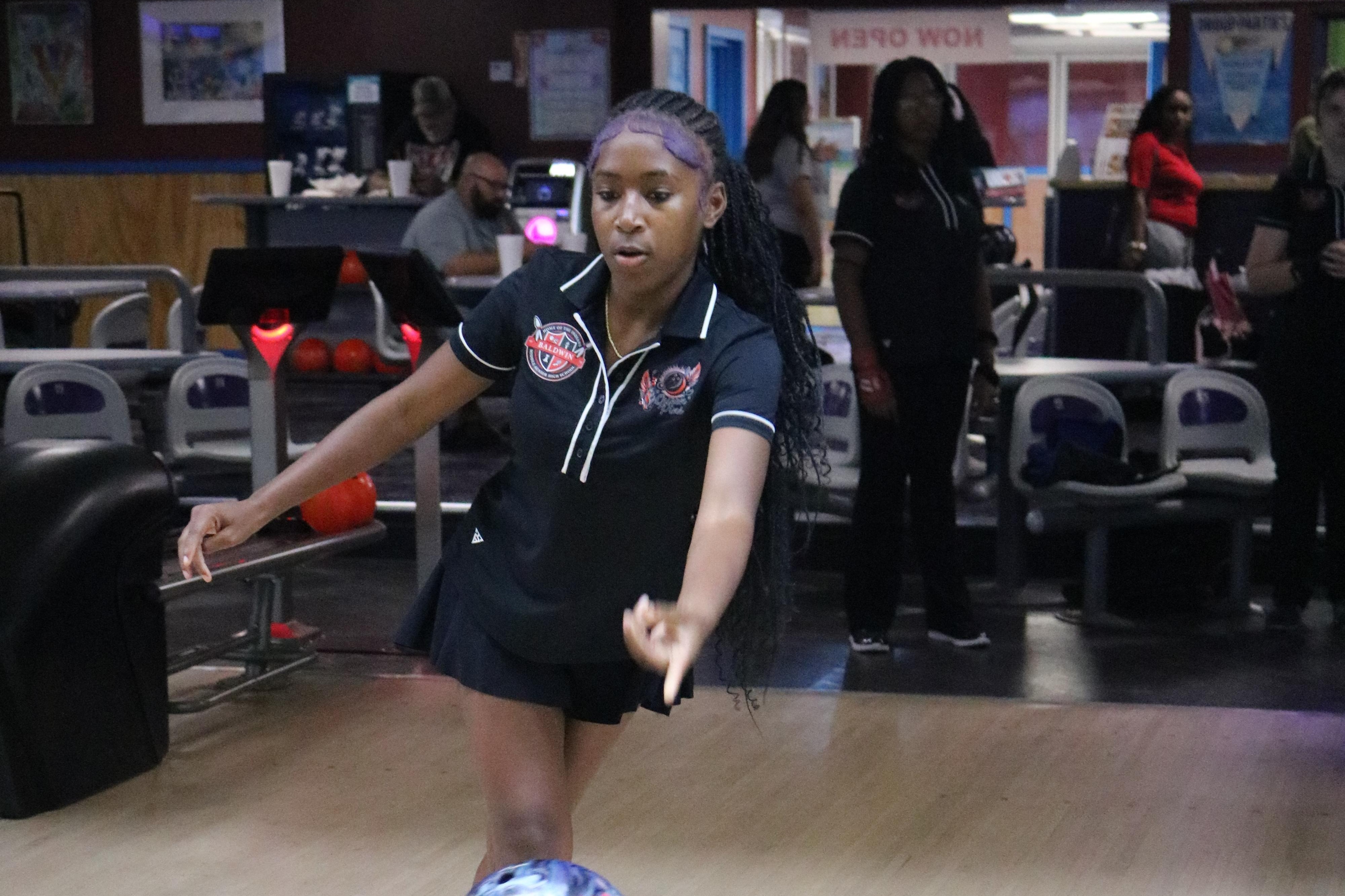 A young person throws a bowling ball down the lane. There are bowling pins, a wooden lane, and spectators.