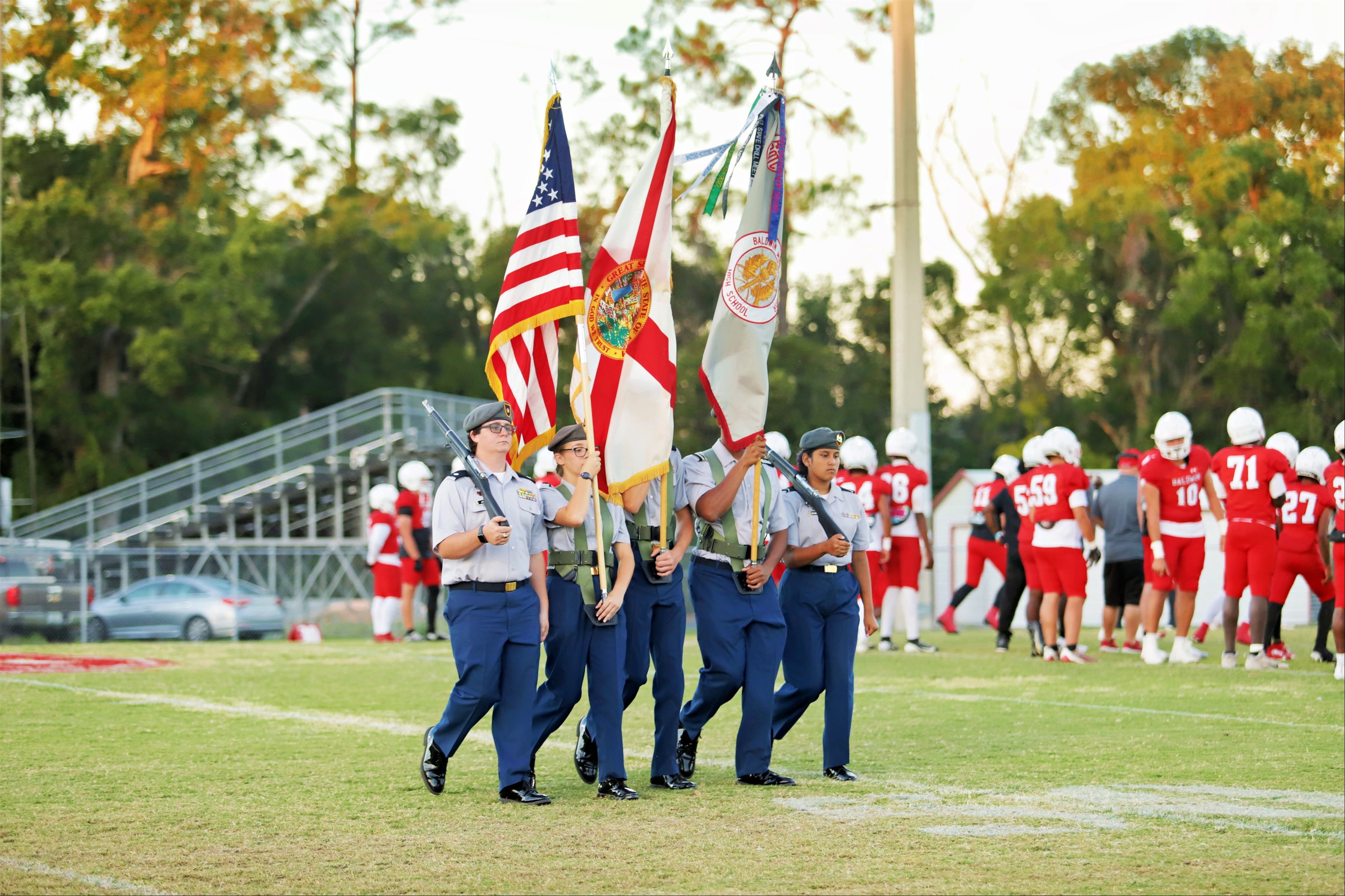 Six individuals in uniform march across a field carrying flags. A football team stands in the background.