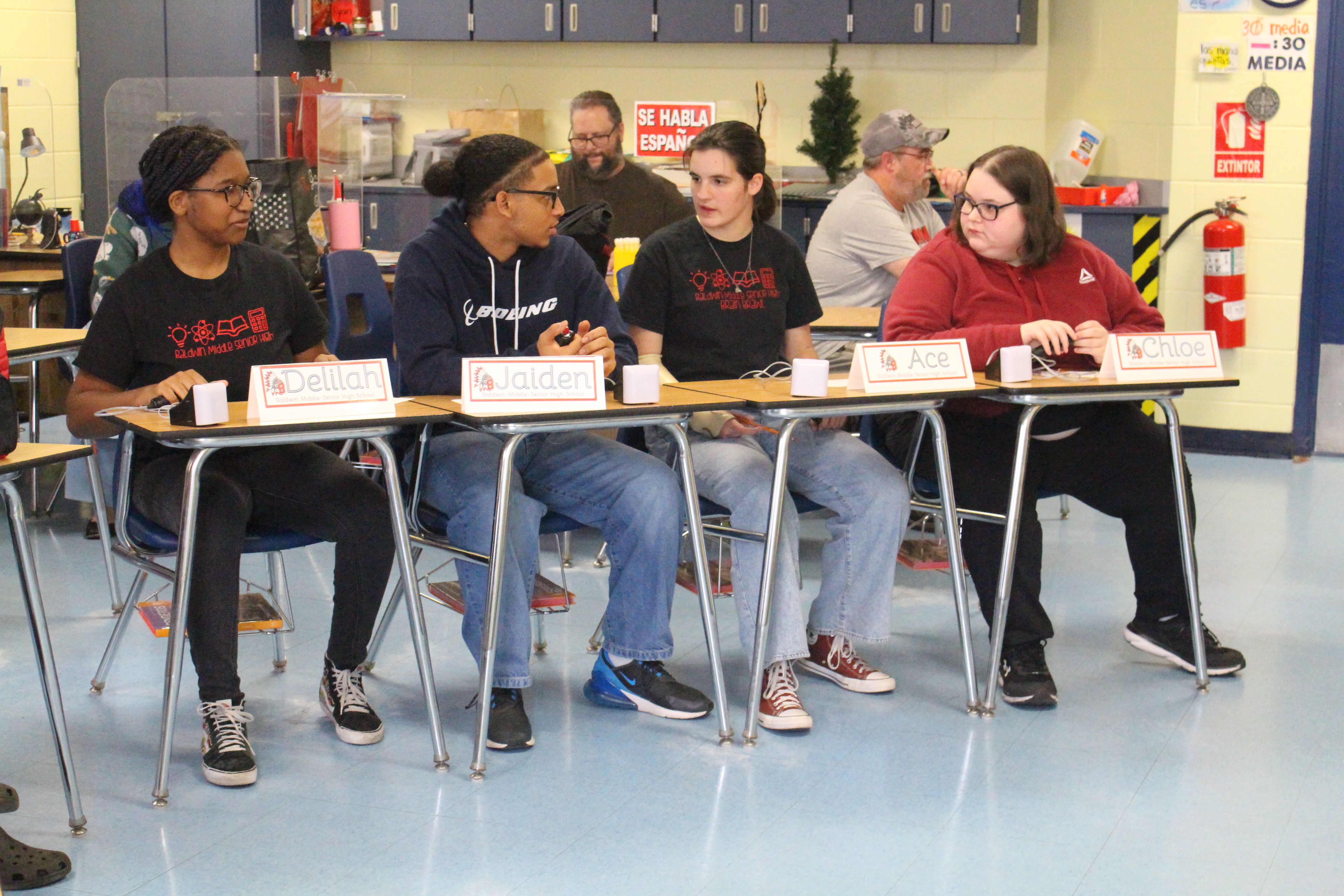 Five individuals seated at a table in a classroom, wearing casual clothes, looking at papers.