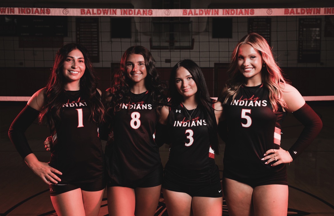 Four female volleyball players in black jerseys pose with numbers 1, 3, 5, and 8. They stand in a gym.
