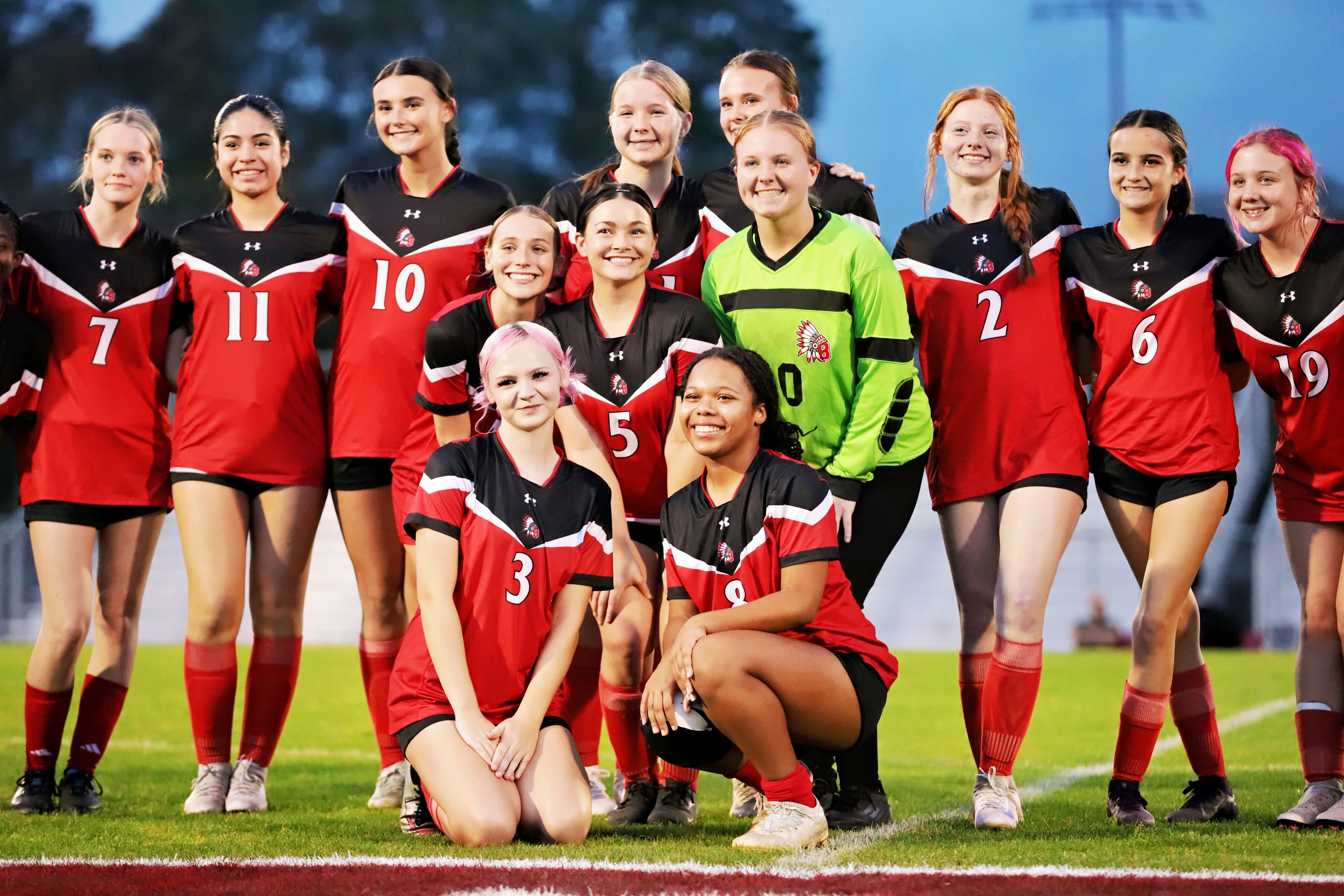 A group of women in red and black jerseys stands on a field. They wear numbered jerseys, and one person crouches.