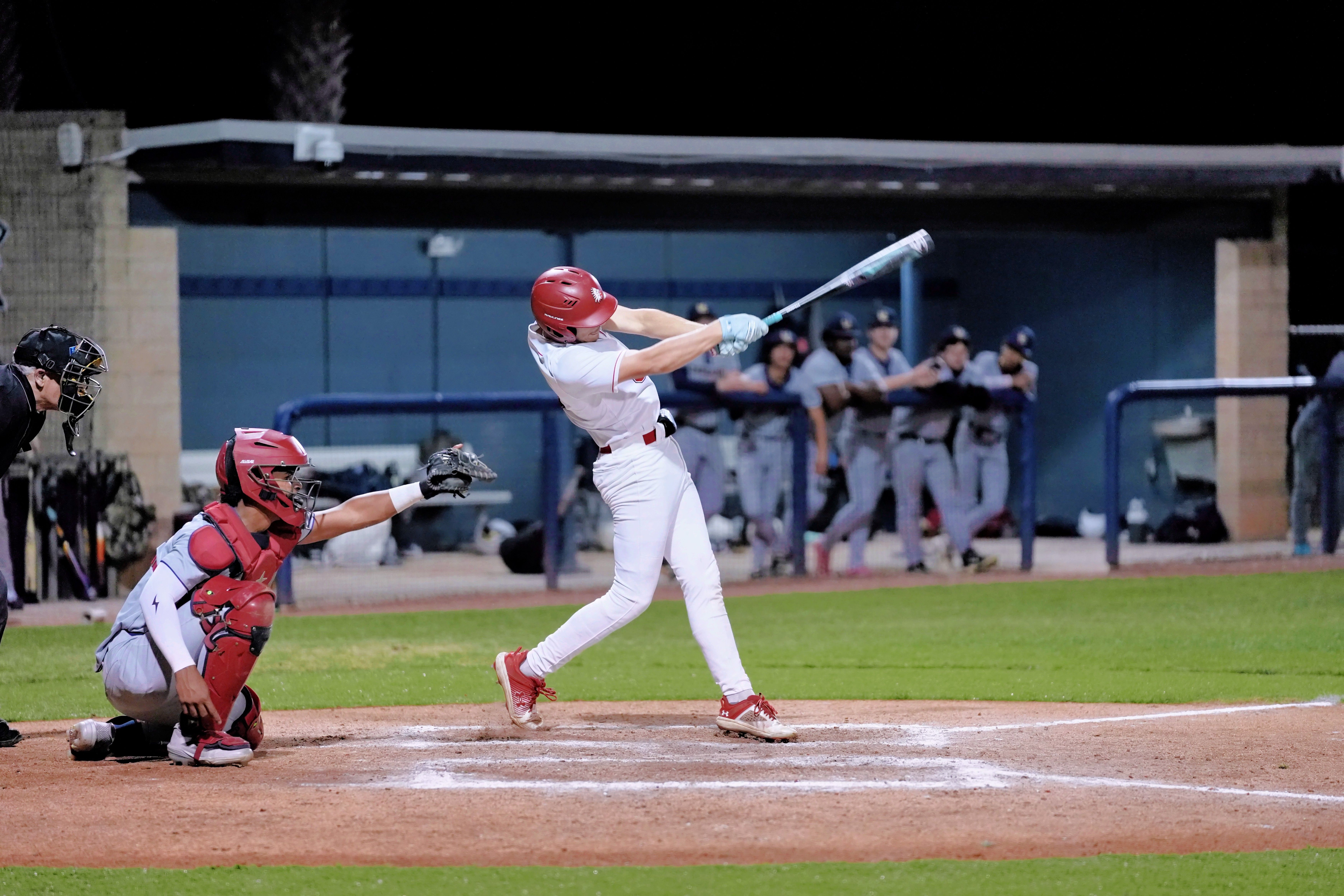 A baseball player swings a bat at a game. A catcher crouches behind home plate.