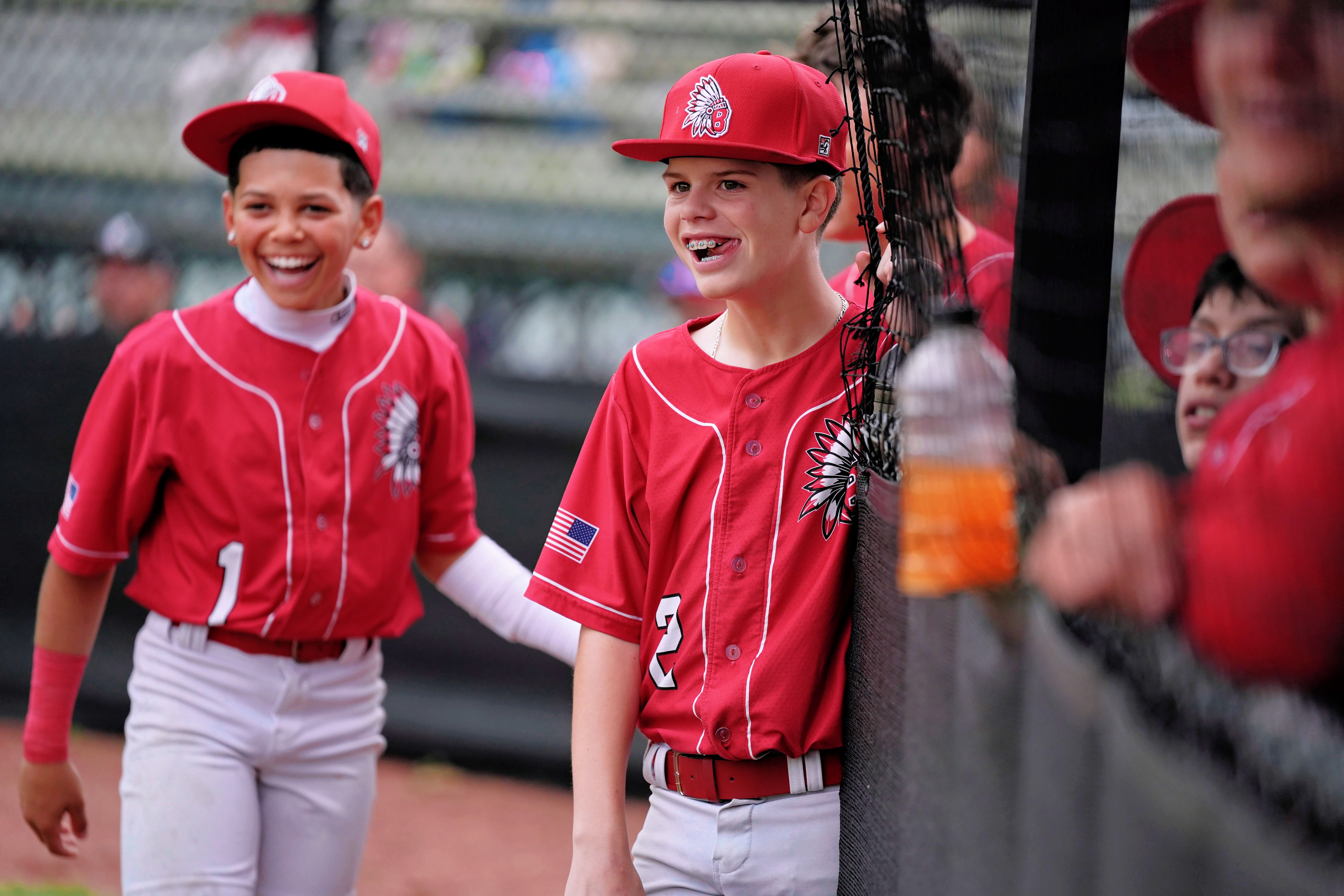 Two children wearing red baseball uniforms, hats, and sneakers walk toward a fence. One has number 1, the other 3.