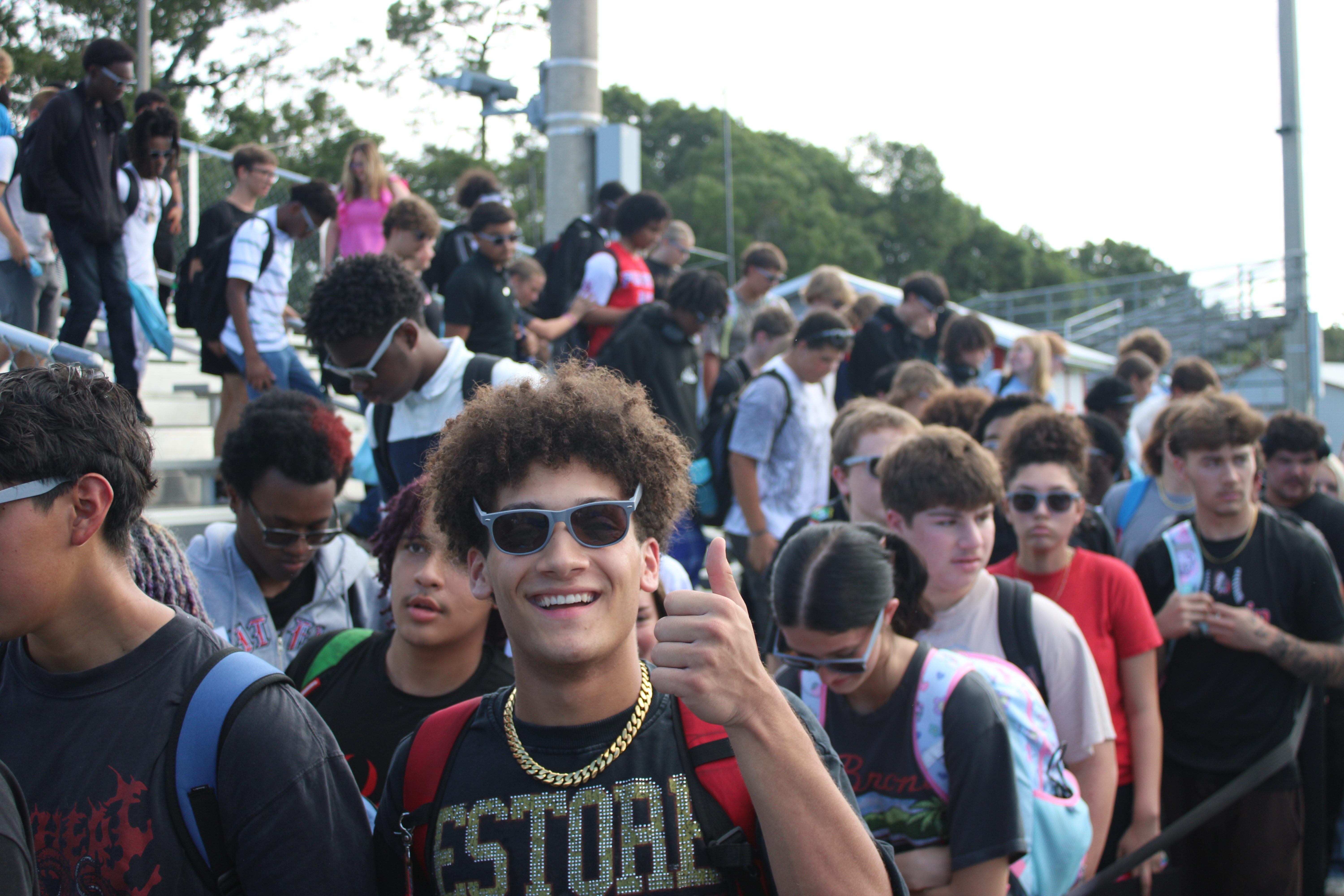 Crowd of students on bleachers with one wearing a gold necklace and sunglasses giving a thumbs-up.