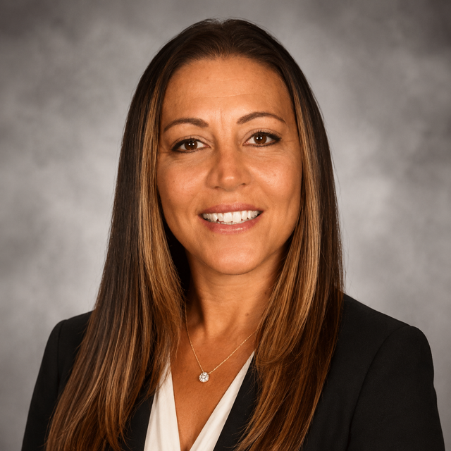 Woman in a black blazer and white shirt, smiling. She has long, wavy brown hair.