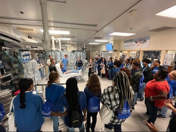 People in blue lab coats and backpacks stand in a line in a room with white walls and fluorescent lights.