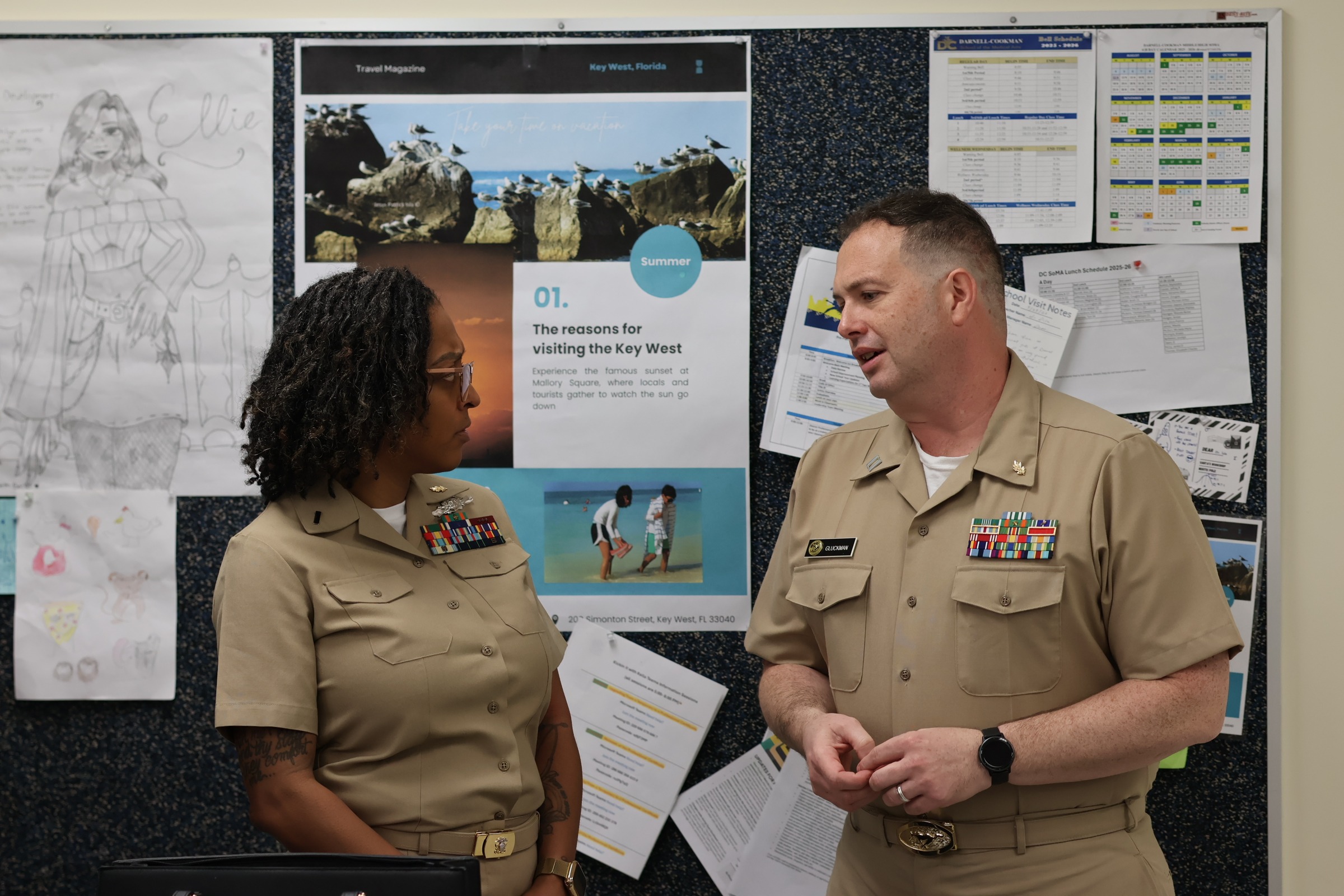 Two individuals in military uniforms stand in front of a bulletin board with papers attached.