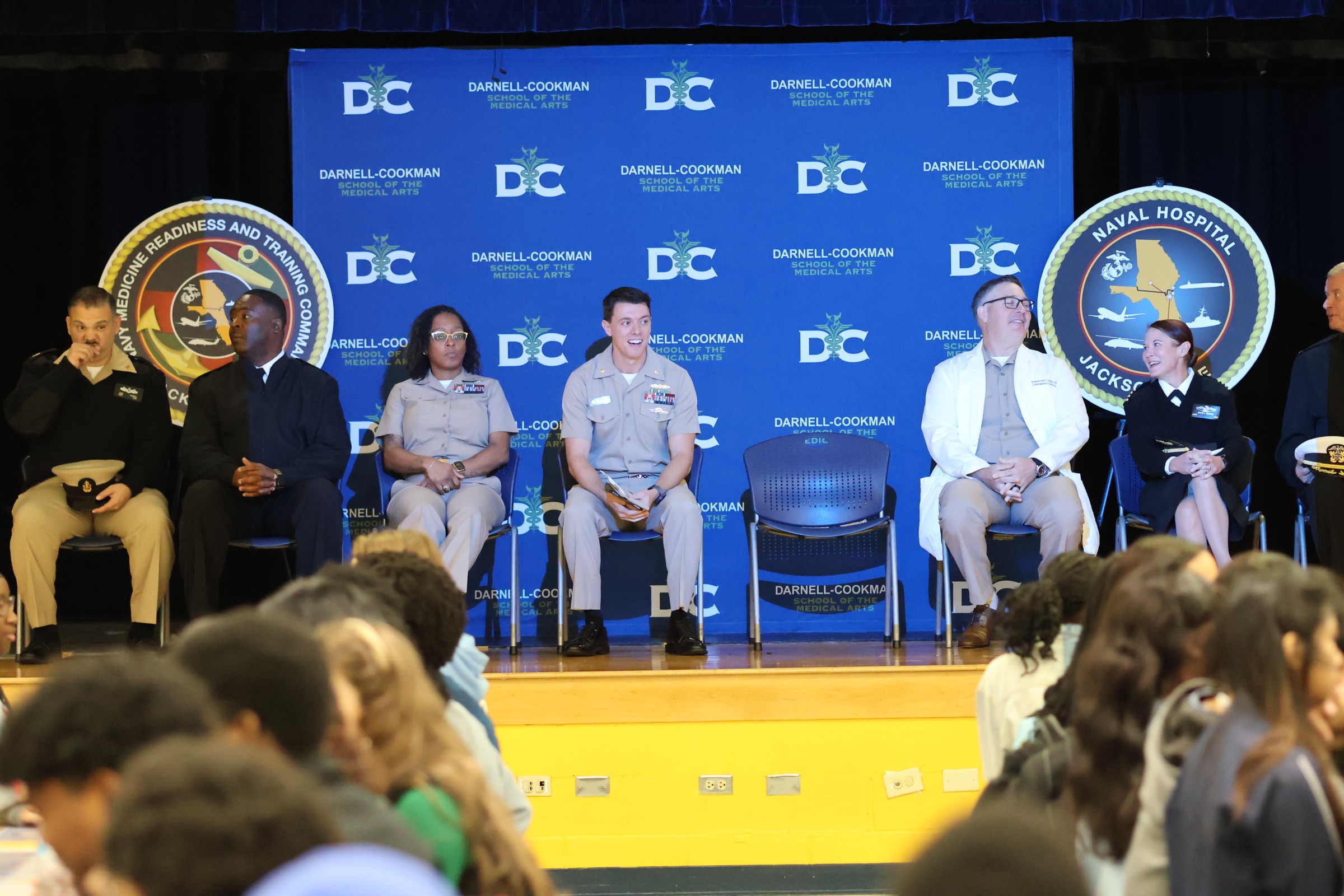 People sit on stage with a backdrop. Three wear uniforms. Two hold papers. One holds a microphone.