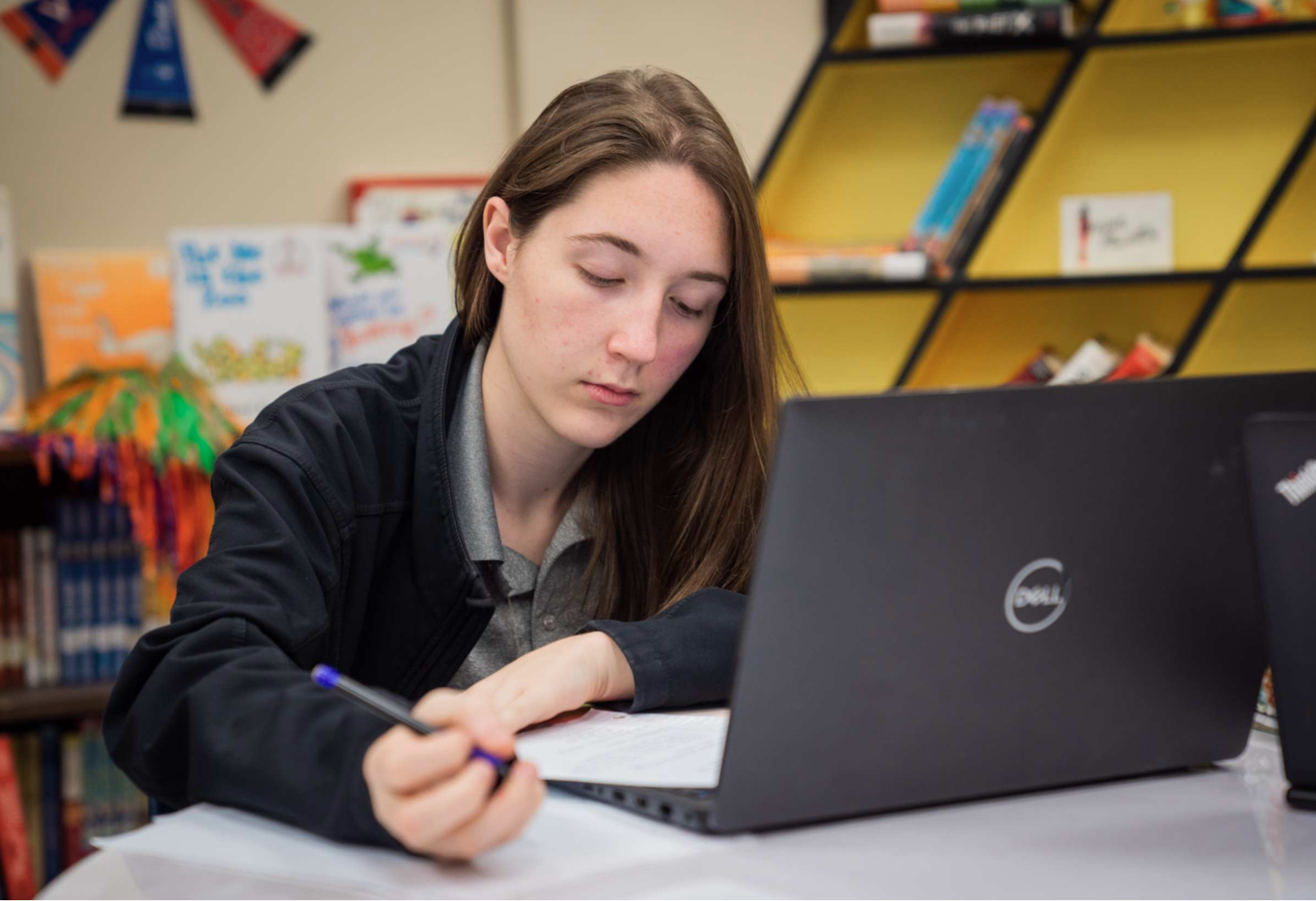 A student with brown hair works on a laptop at a desk in a classroom with bookshelves.