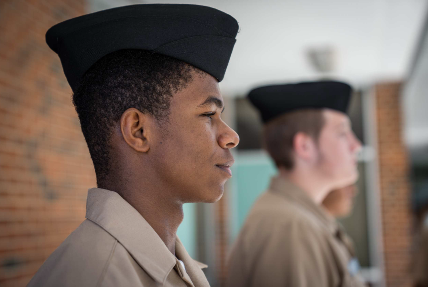 Two individuals in military uniforms and black hats stand in a corridor. One person faces forward, and another looks to the side. A brick wall is behind them.