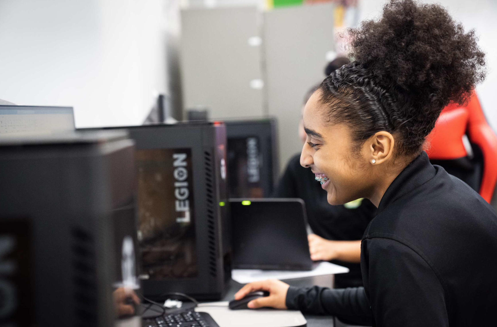 A young woman sits at a computer, smiling, wearing a black shirt. Behind her, another person works at a computer.