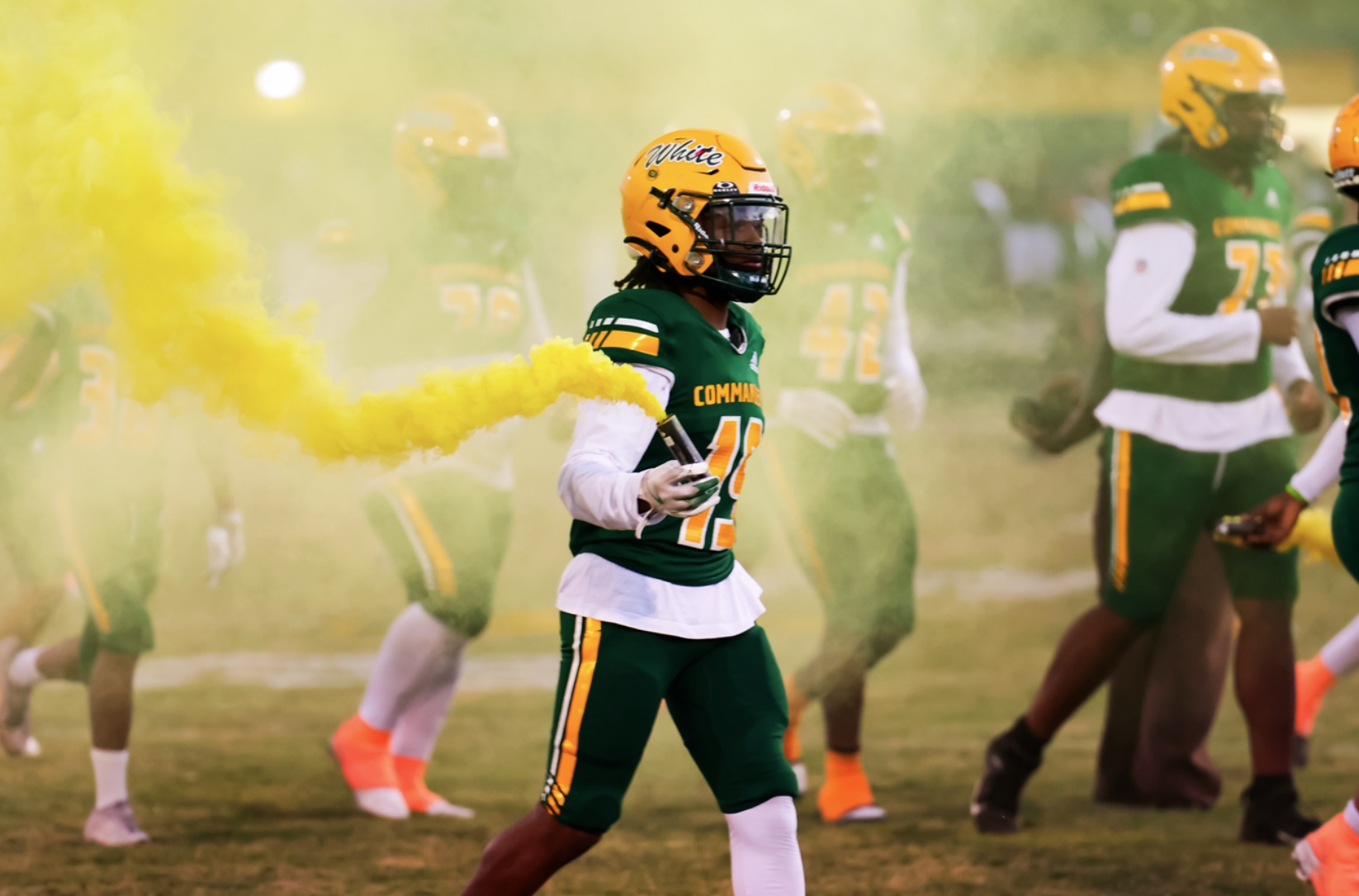Players in green and white uniforms, with one player holding yellow smoke, run on a grassy field.