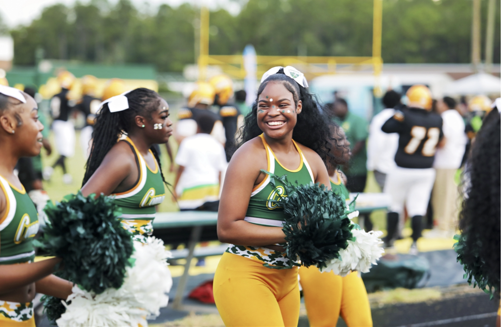 Cheerleaders in green and yellow uniforms stand on the field, smiling, with a crowd of people in the background.