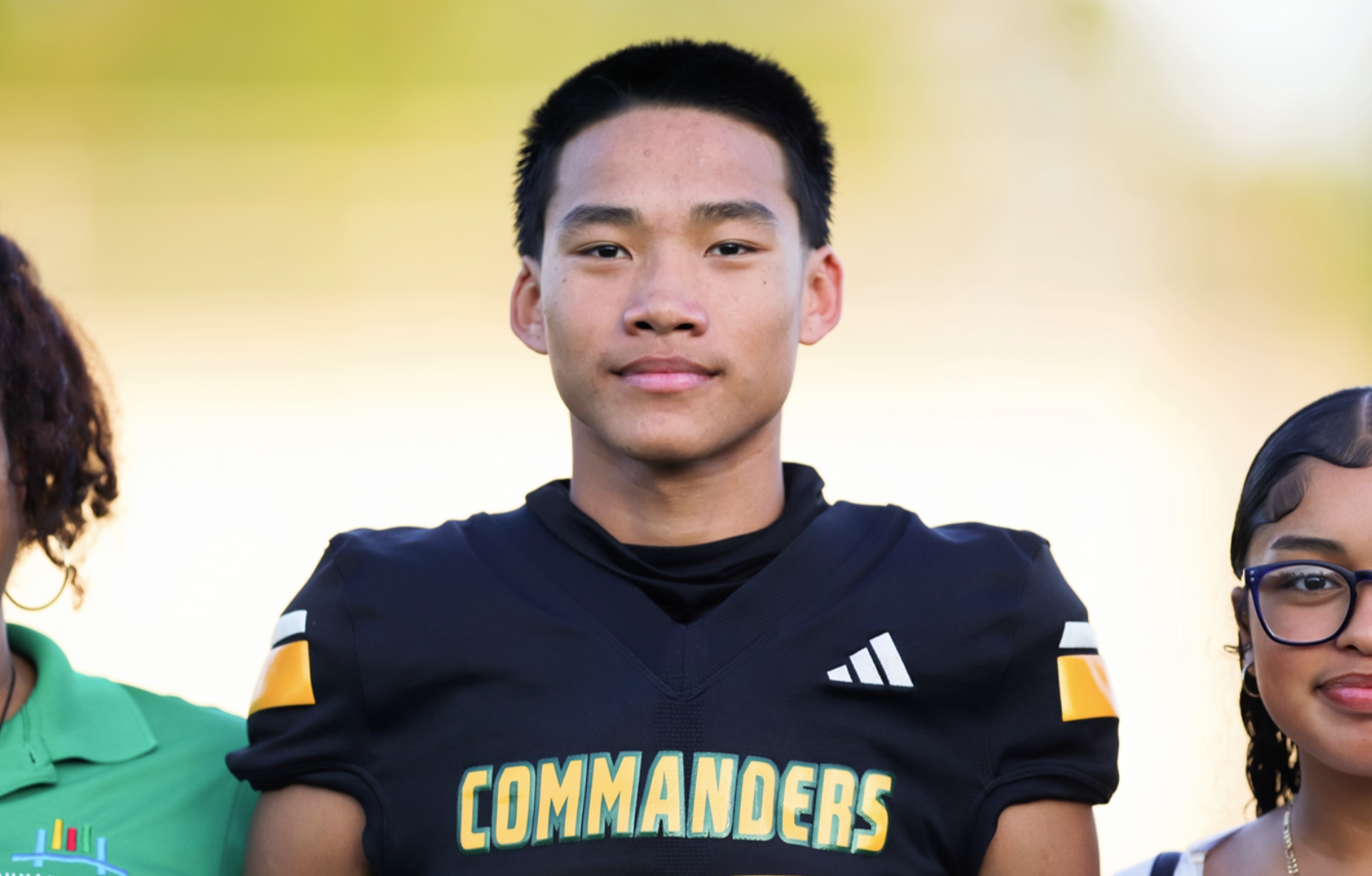 A young man in a black football jersey with "COMMANDERS" on it stands between two others. The background is blurred.