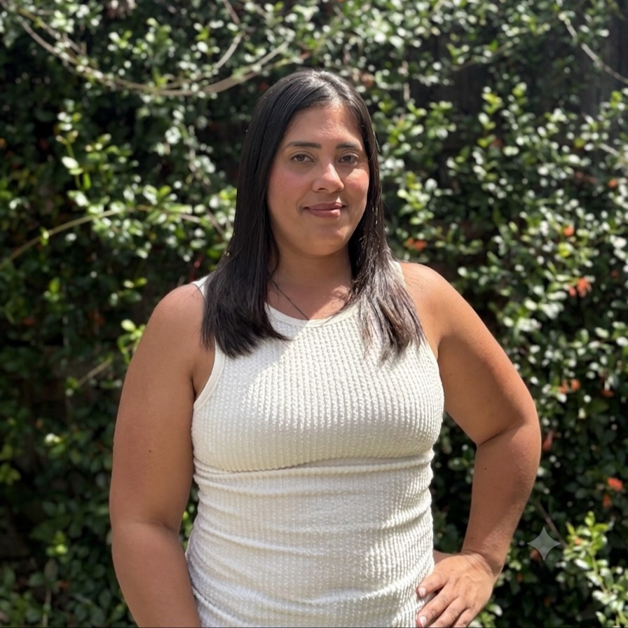 Person with dark hair wearing a white sleeveless top, standing before a green hedge with small red flowers.
