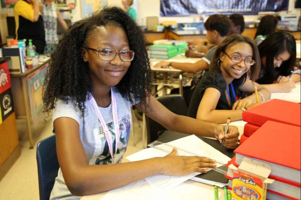 Students in a classroom. A girl with glasses smiles at the camera. Another student sits nearby, writing.