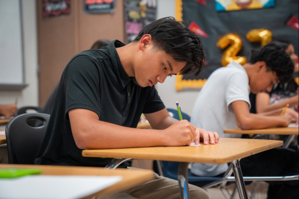A classroom with students seated at desks, one focused on writing, others seated, some writing, a bulletin board behind them.