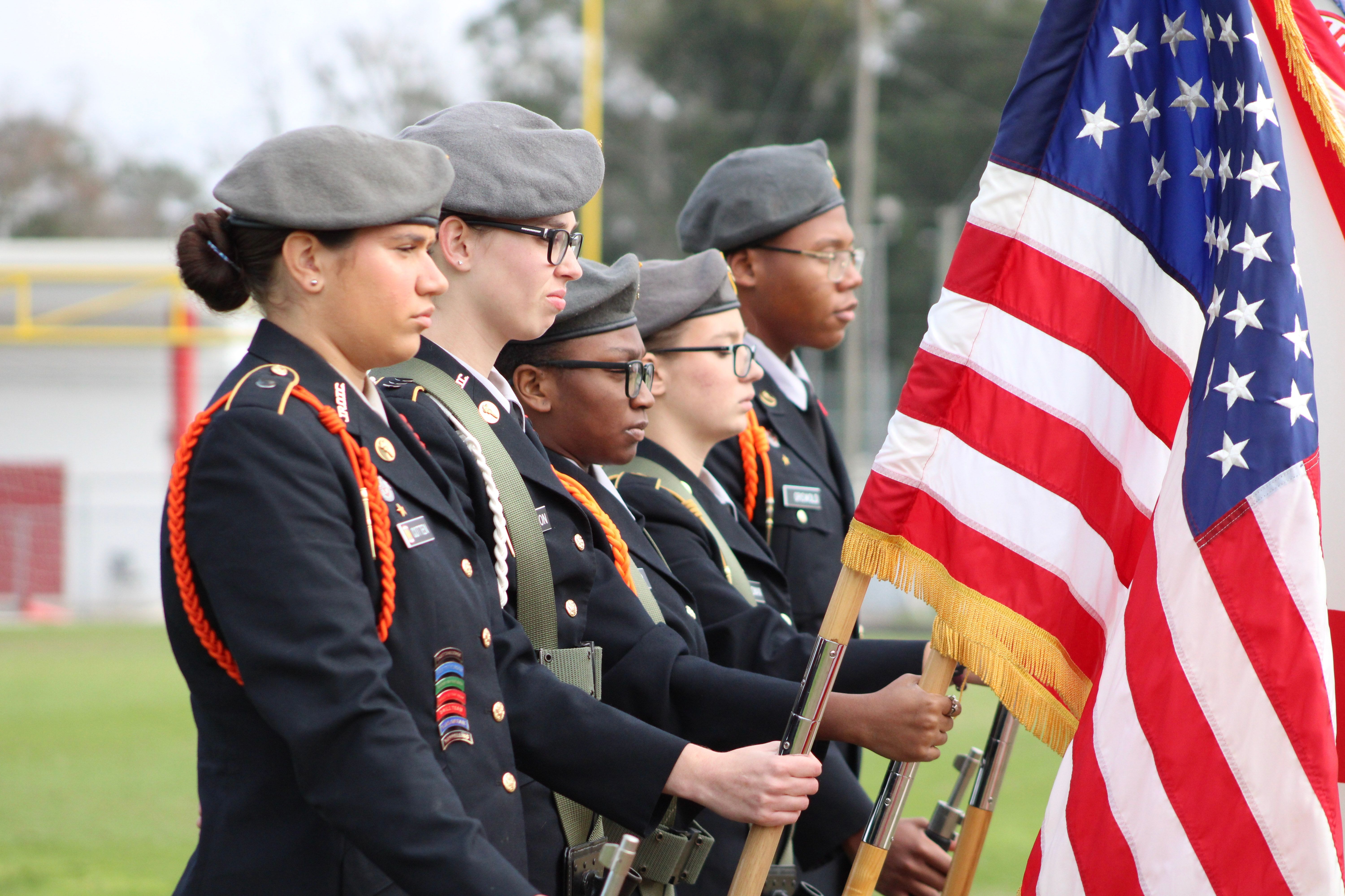 Five military cadets, three women and two men, holding American flags and rifles. They are in uniform and standing in a field.
