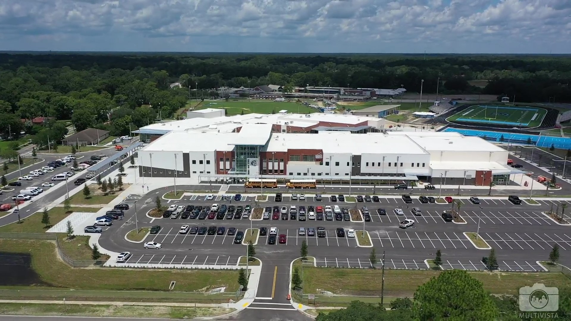Aerial view of a modern, large building with a parking lot full of cars. Trees and a field are visible in the background.