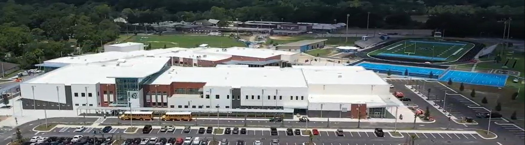 Aerial view of a large white building with a brown entrance, a parking lot, and a sports field.