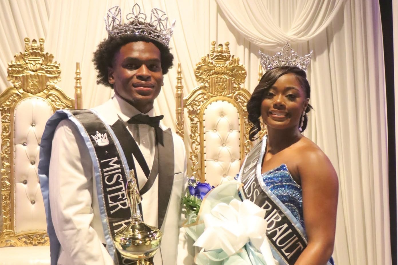 Man and woman standing, holding sashes and trophies, smiling for a photo. Gold chairs behind them.