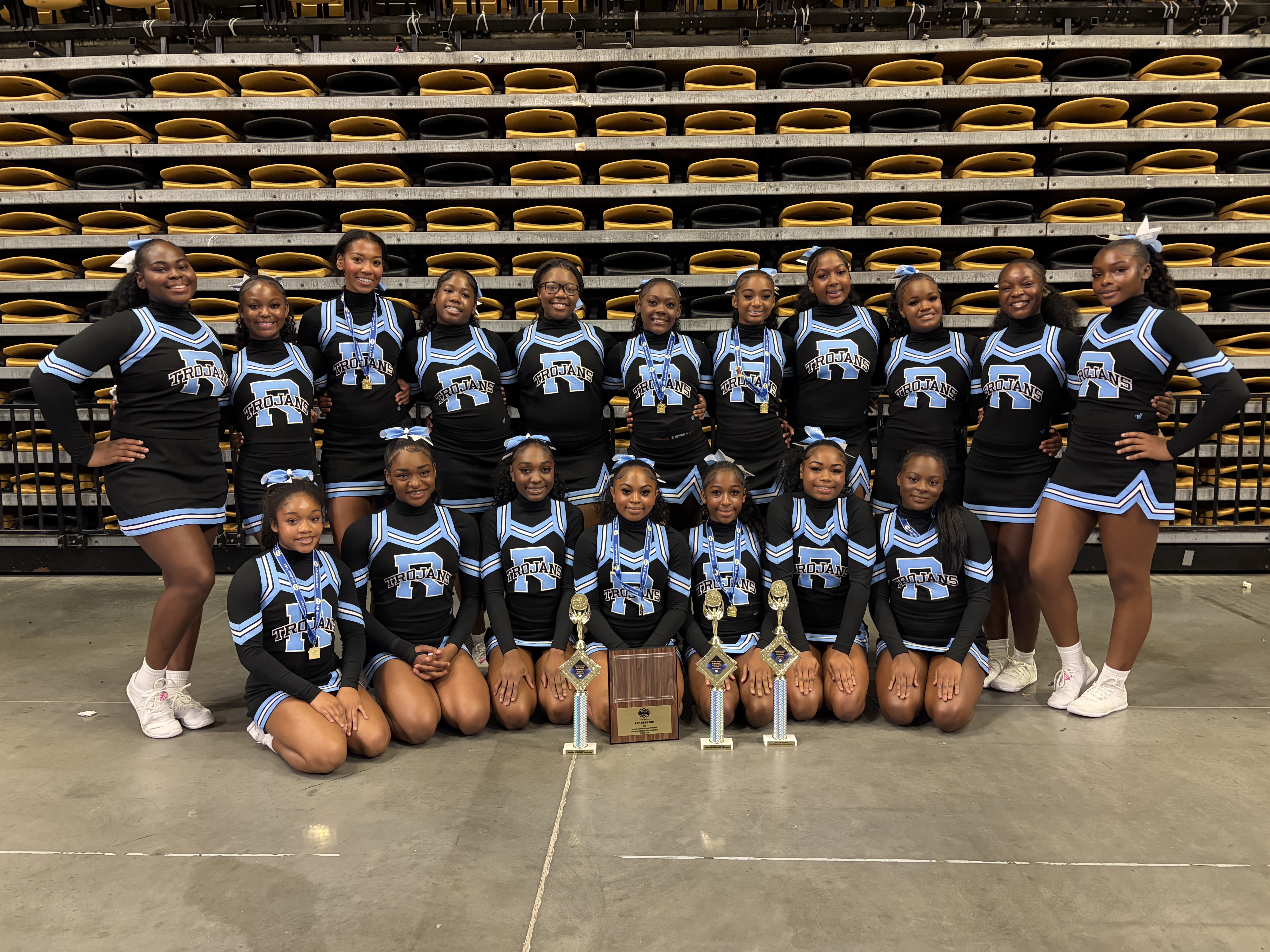 A cheerleading team in matching uniforms poses for a photo with trophies in front of empty stadium seating.