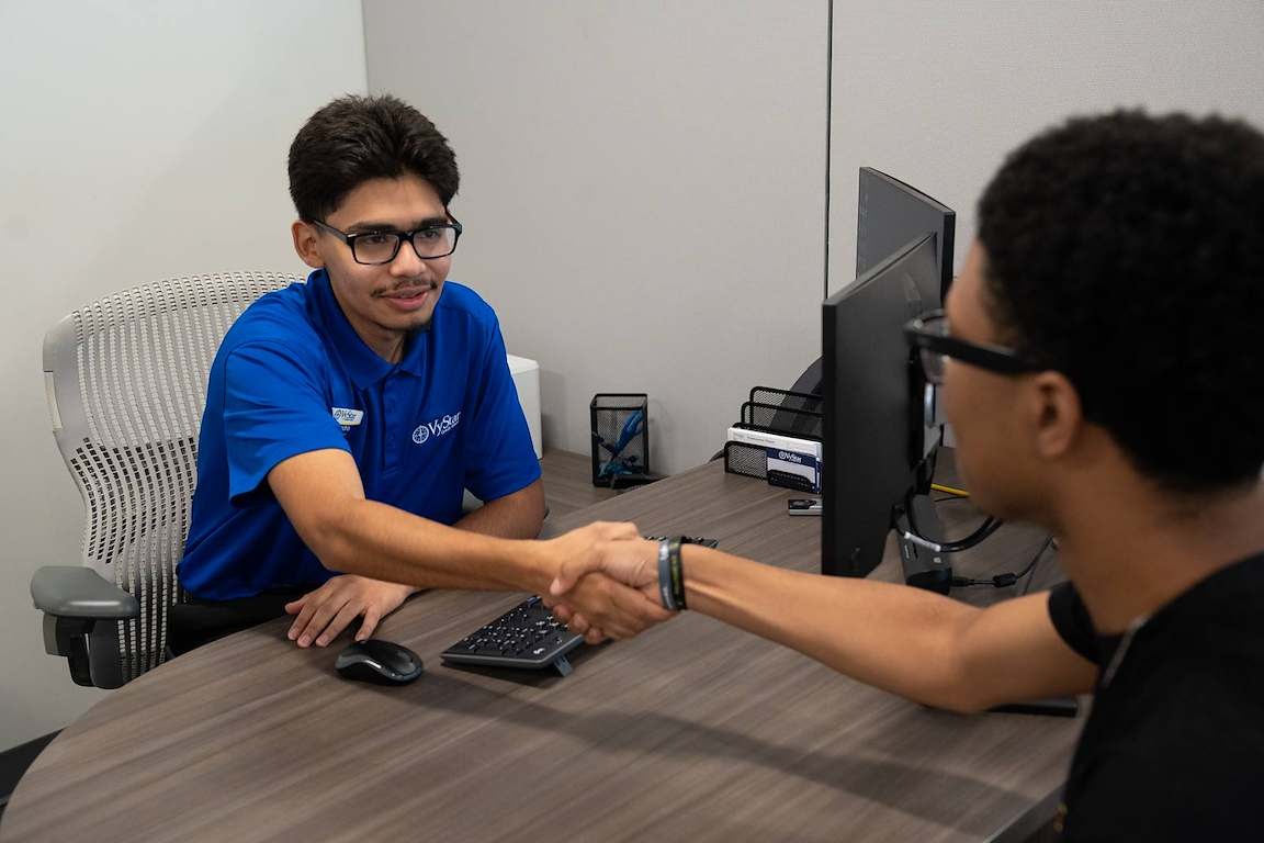 Two people sit at a desk, shaking hands. One wears a blue shirt and glasses, the other a black shirt.